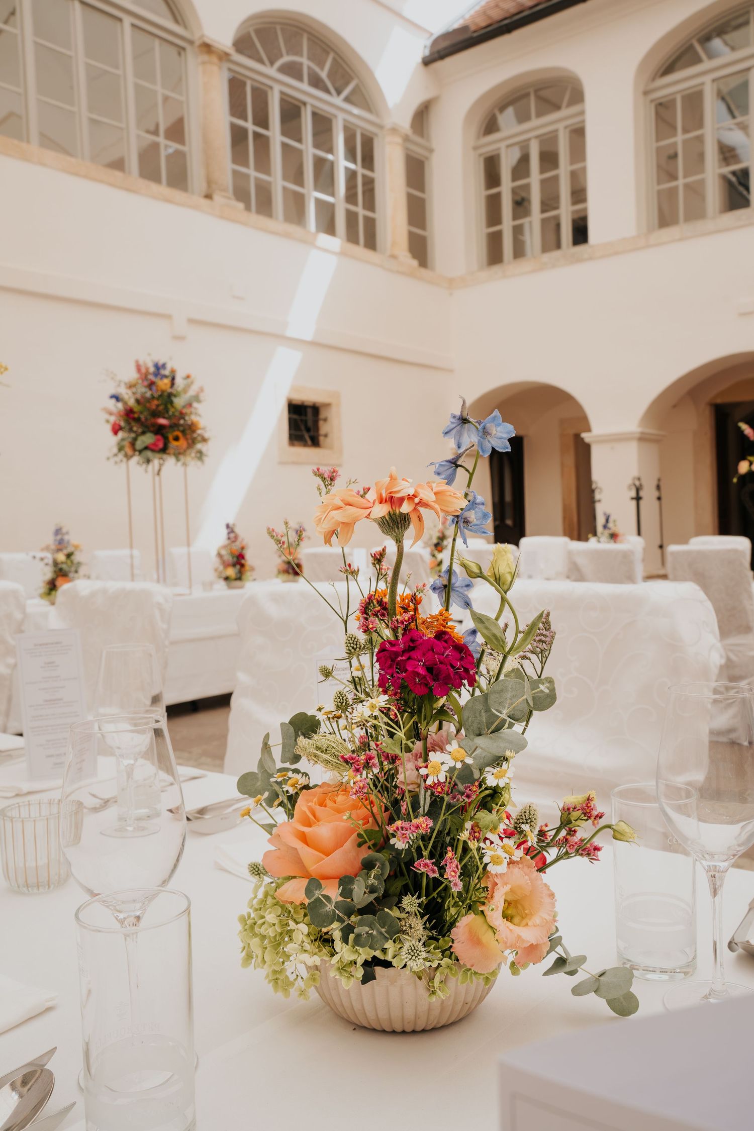 Elegant wedding reception centerpiece with peach roses, red dahlias, and wildflowers in a courtyard venue with white draped tables.