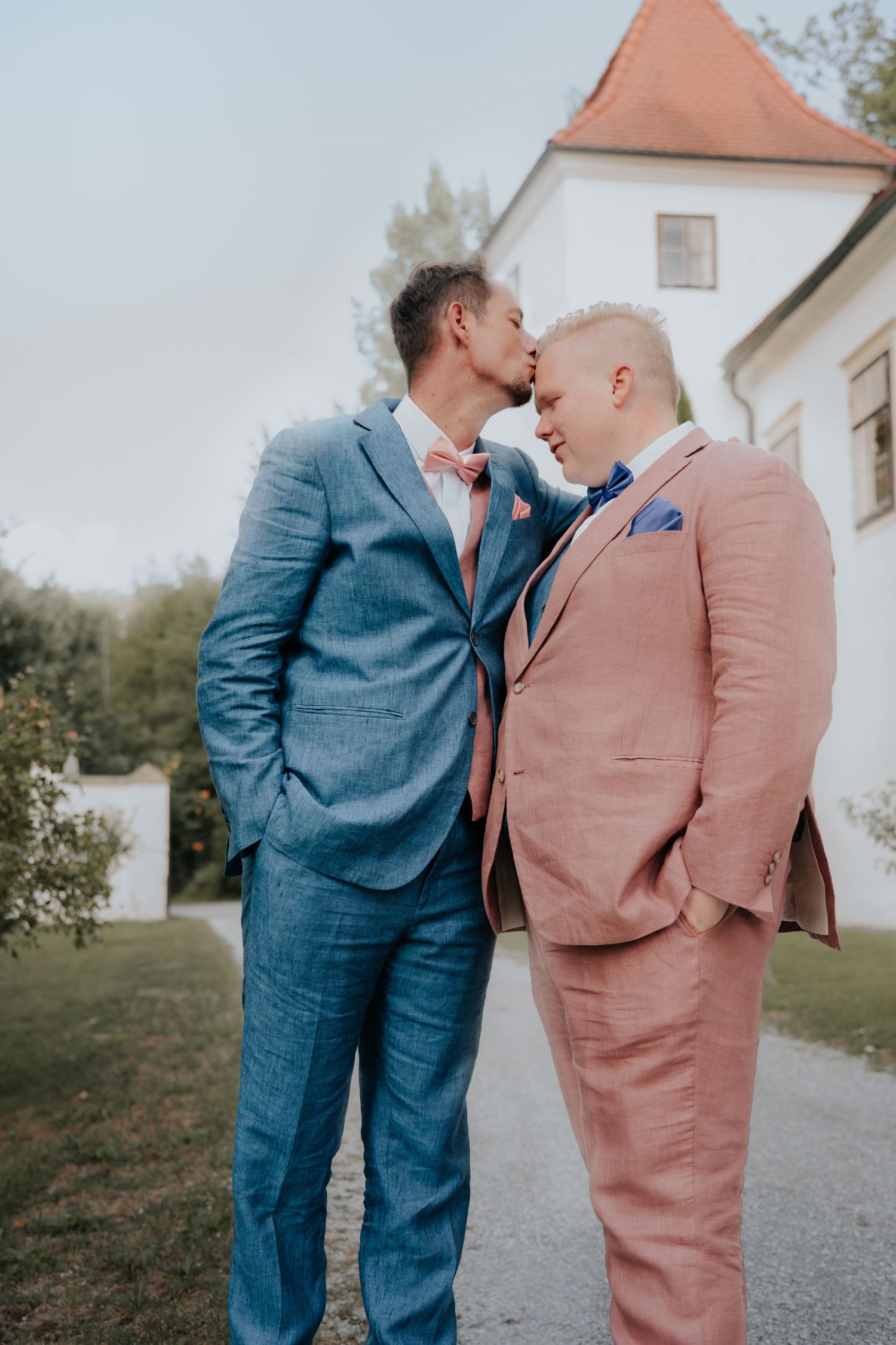 Two grooms in blue and pink suits share a tender kiss outdoors near a white building on their wedding day.