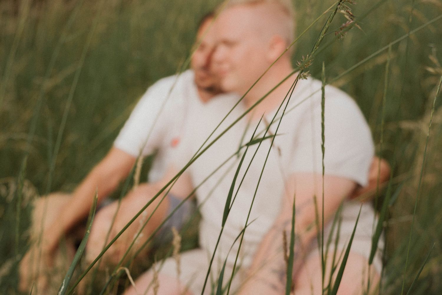 Couple sitting together in a lush green field, surrounded by tall grass in soft natural light.