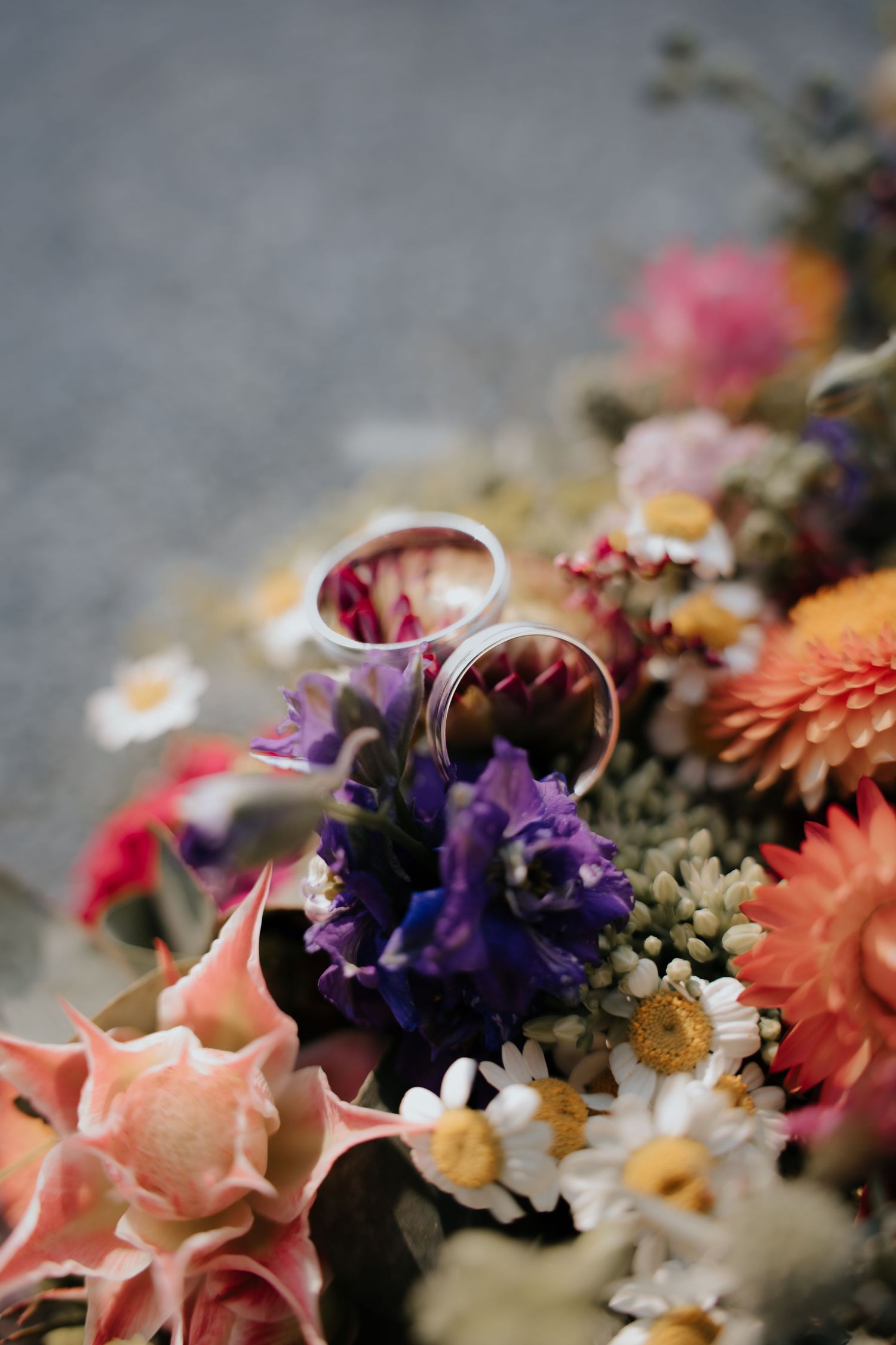 Two silver wedding rings nestled among colorful wildflowers including purple, orange, and white blooms in a bridal bouquet.