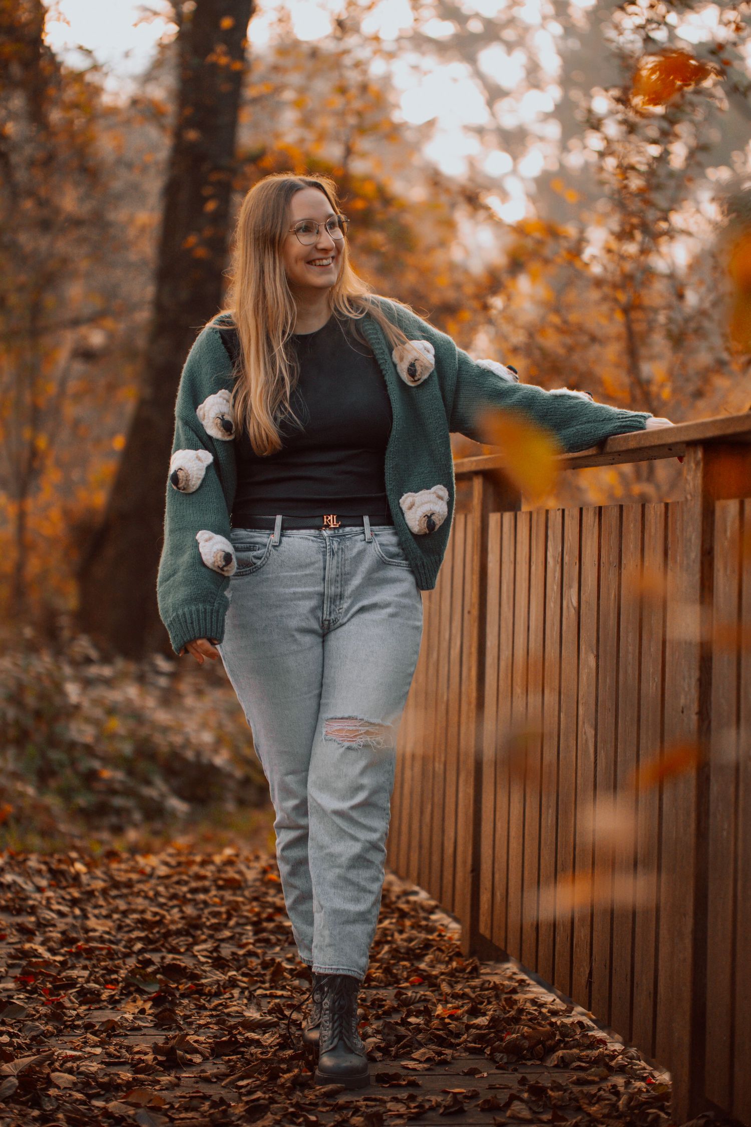 Woman in green cardigan and jeans leaning on wooden fence surrounded by autumn foliage in a forest park.