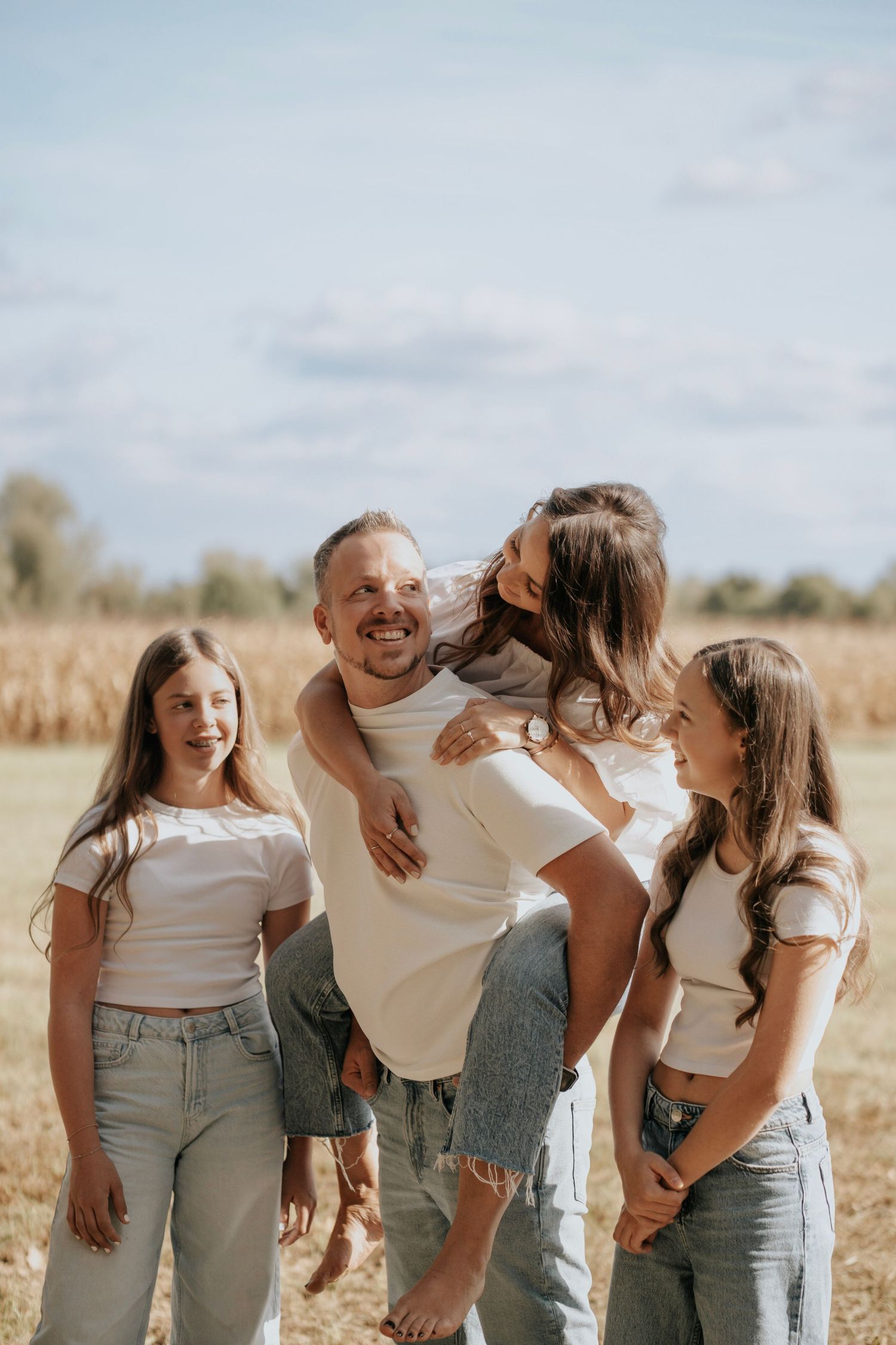Happy family of four enjoying a sunny day in a golden field, father giving piggyback ride to daughter.