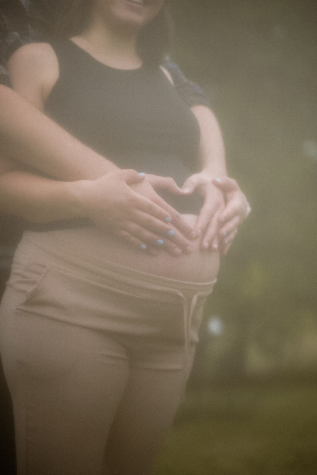 Couple forming heart shape with hands on pregnant belly outdoors in soft bokeh light.