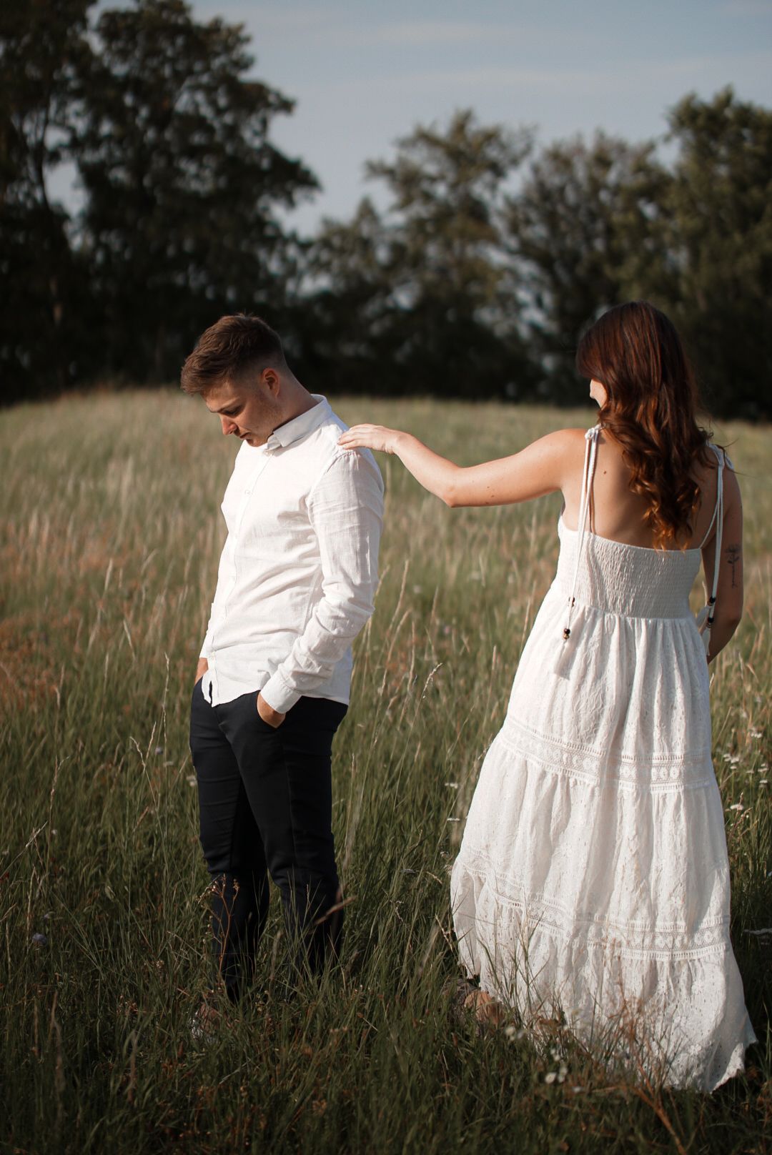 Couple in white outfits walking through a green field, woman in flowing dress touching man's shoulder.