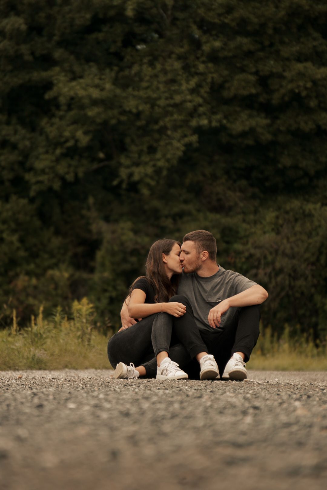 A couple sits on a gravel path kissing, dressed in casual black outfits with white sneakers, surrounded by lush green trees.