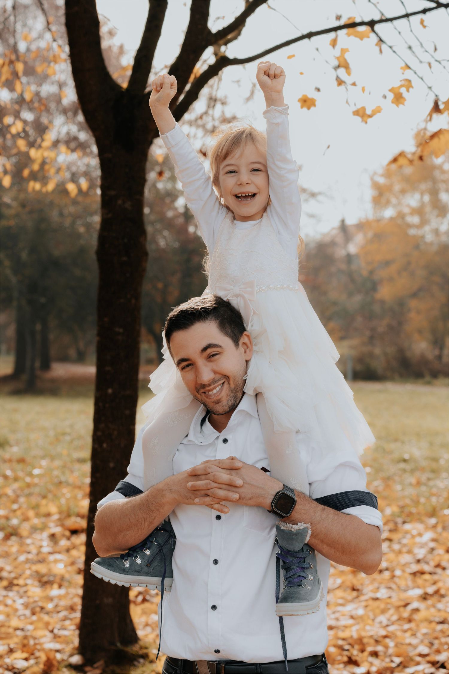 A smiling man carries a joyful little girl on his shoulders in an autumn park, both wearing white outfits.
