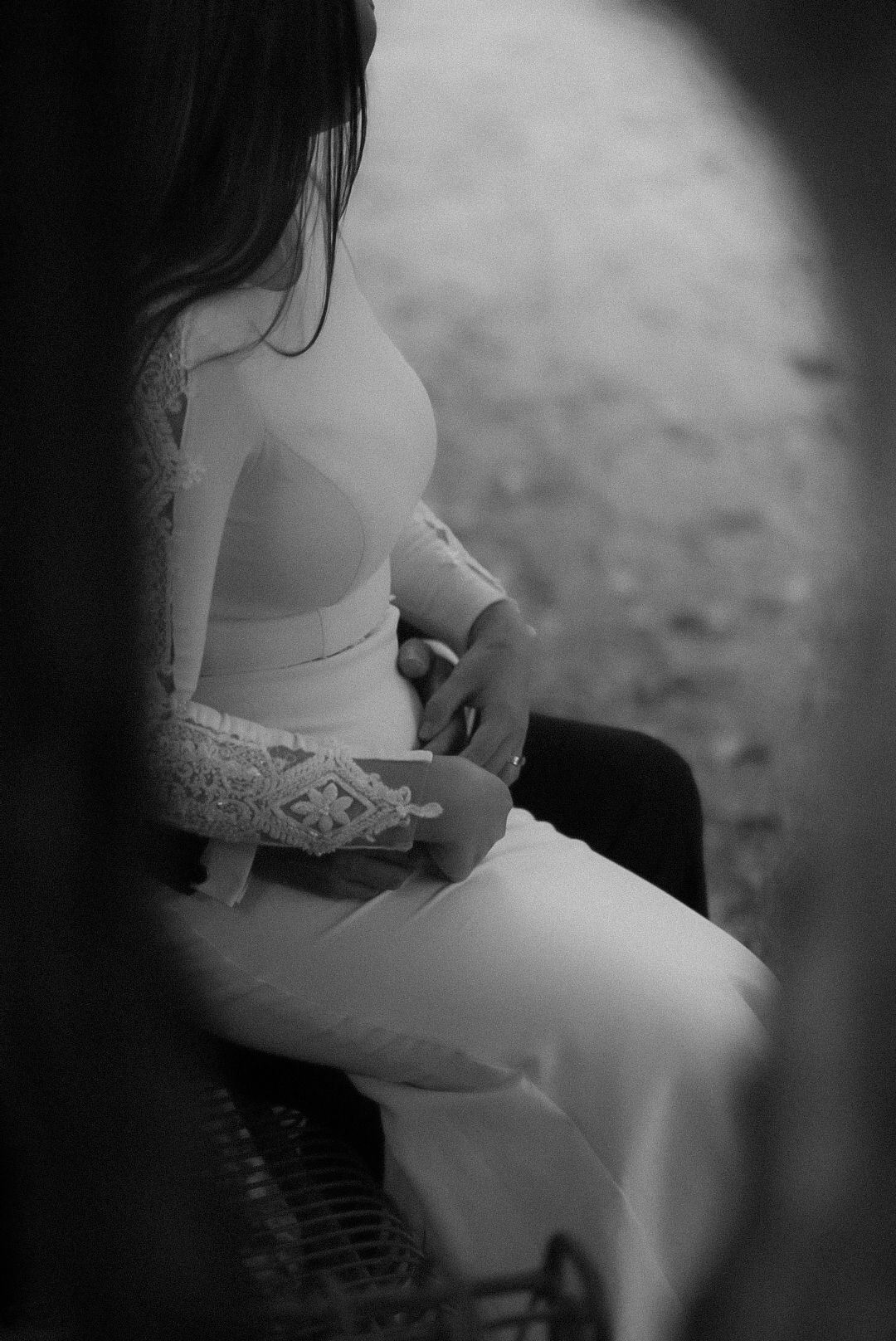Black and white photo of a pregnant woman in a white lace dress sitting gracefully, cradling her baby bump.