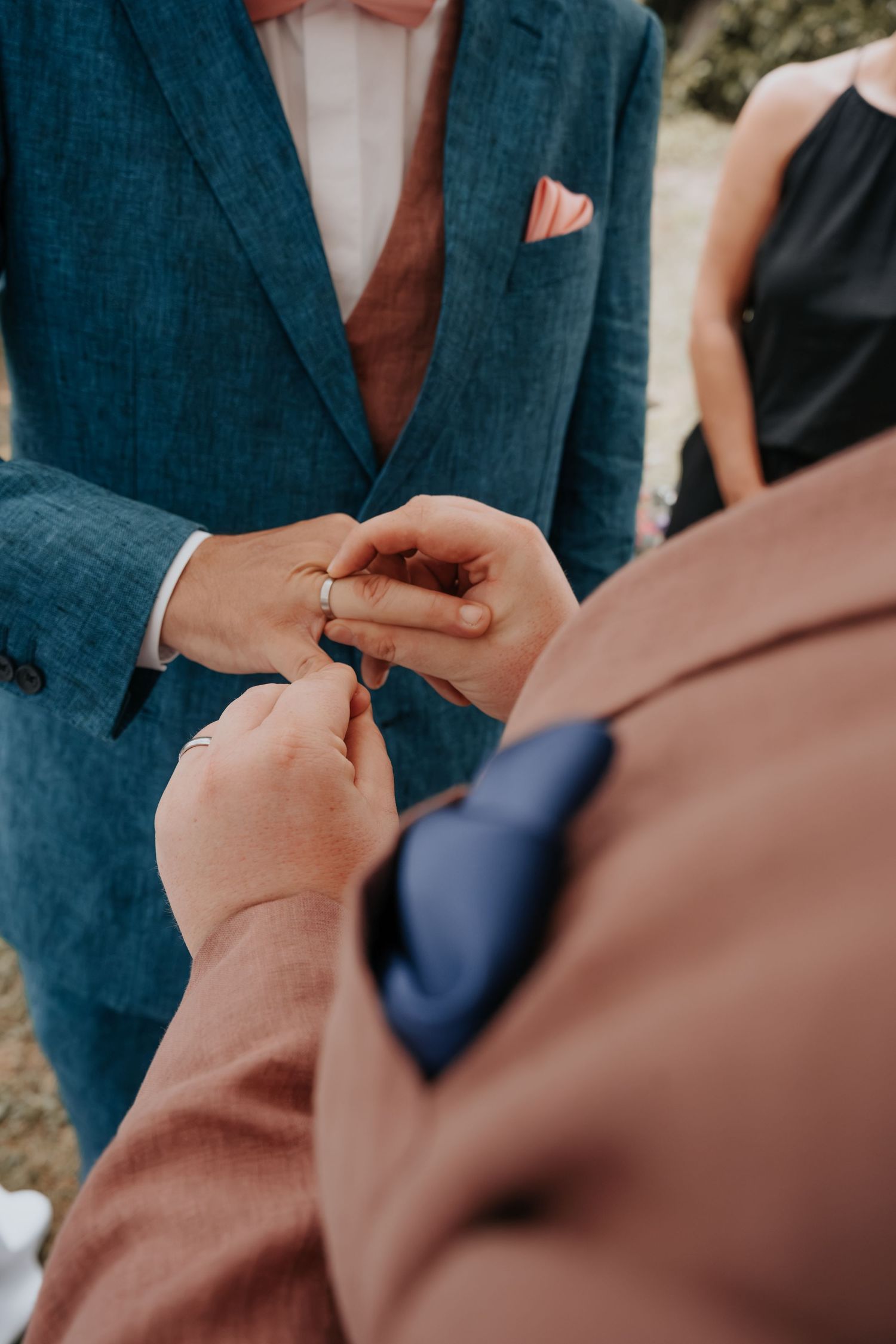 Two grooms exchange wedding rings during ceremony, one wearing teal suit with pink pocket square, other in mauve suit with navy tie.