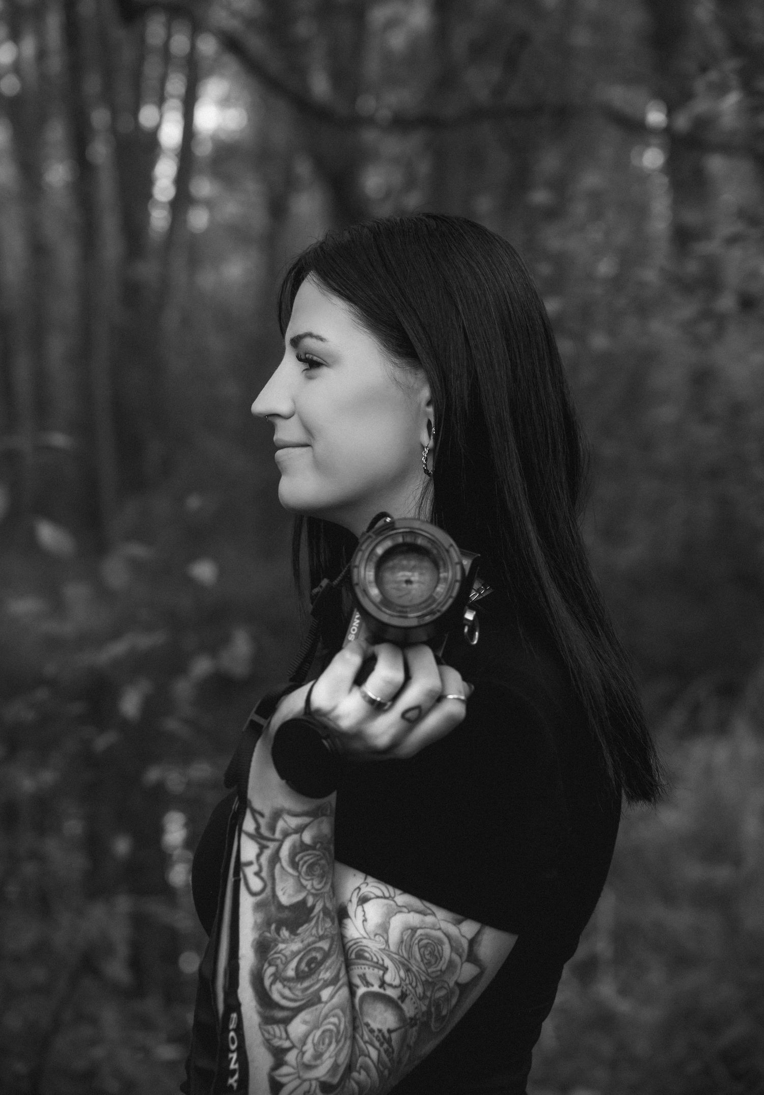 Black and white portrait showing artistic tattoo details in a moody forest setting.