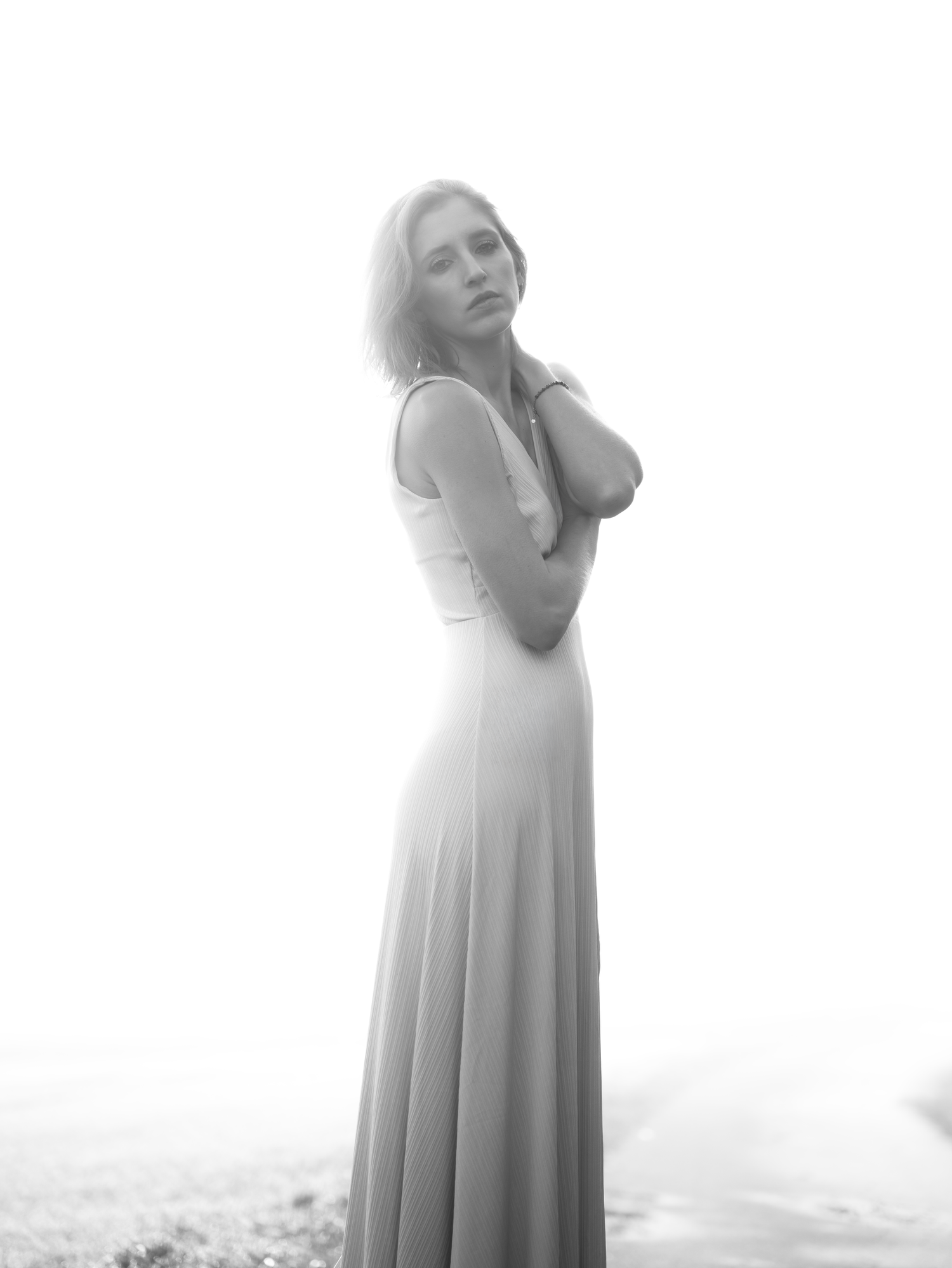 Black and white photo of a woman in a long flowing dress, arms crossed, gazing over her shoulder in a misty outdoor setting.
