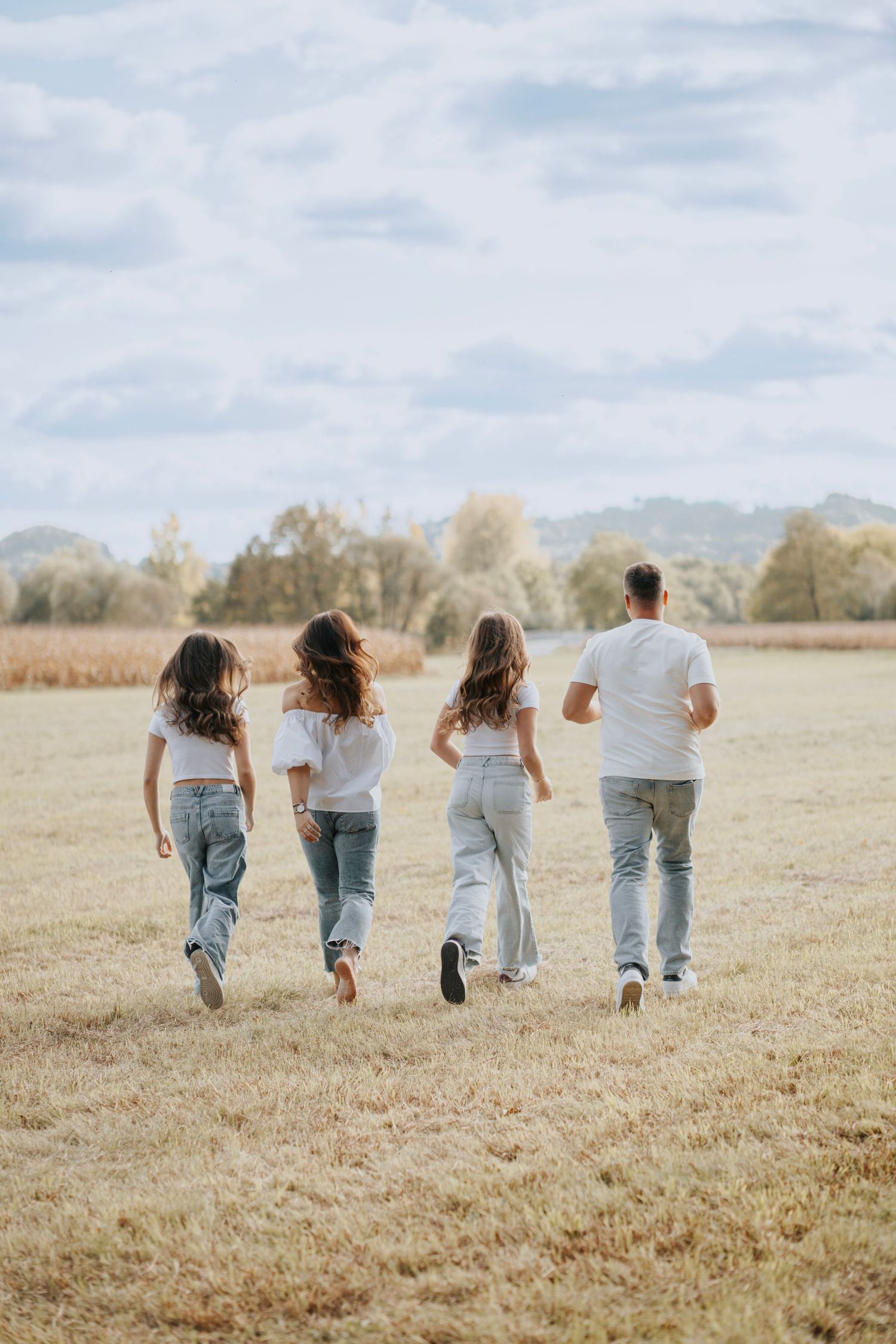 Family of four in white tops and jeans walking through an open field on a sunny day.