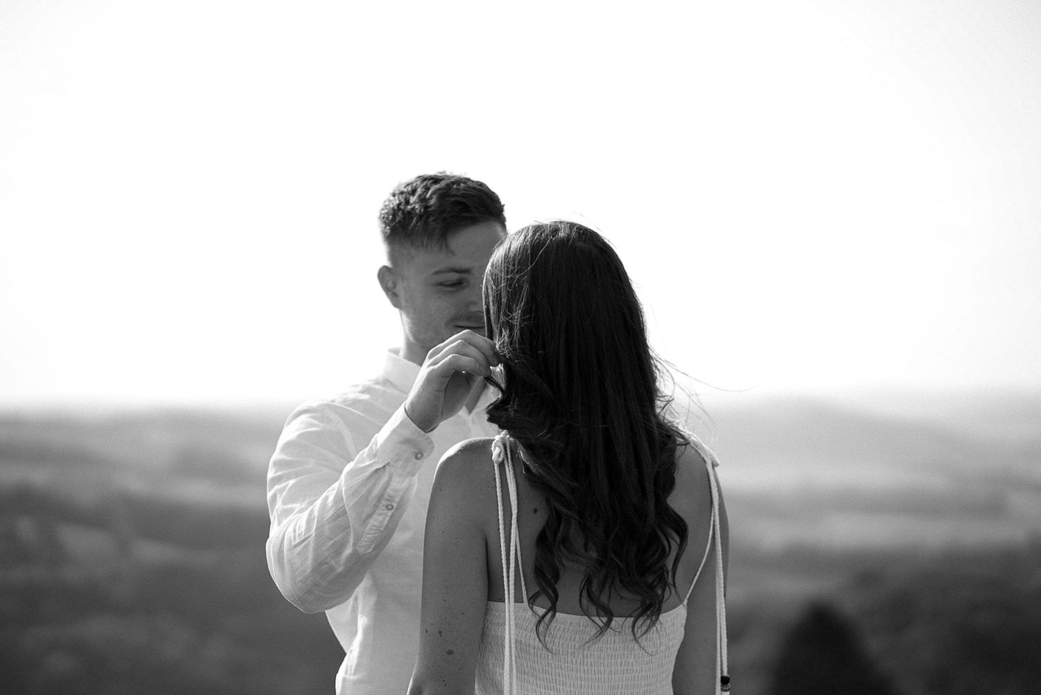 Black and white photo of a couple sharing a romantic moment outdoors with scenic countryside views in the background.