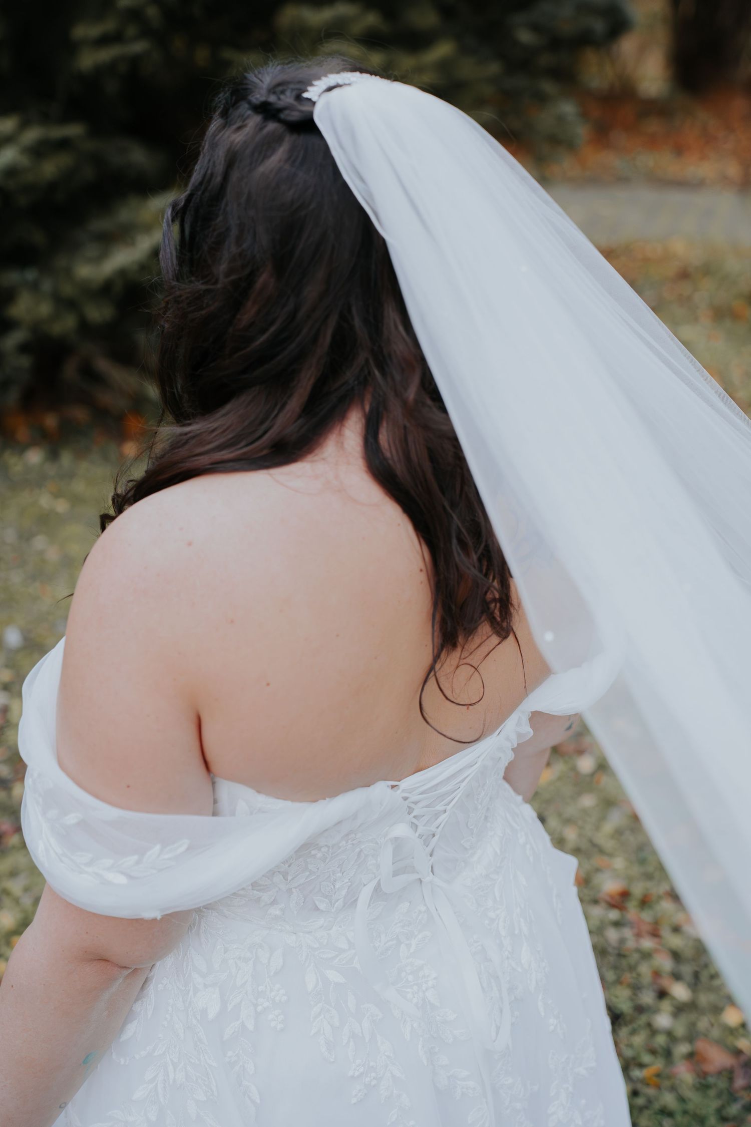 Bride in off-shoulder lace wedding dress and white veil, viewed from behind, with dark hair outdoors.