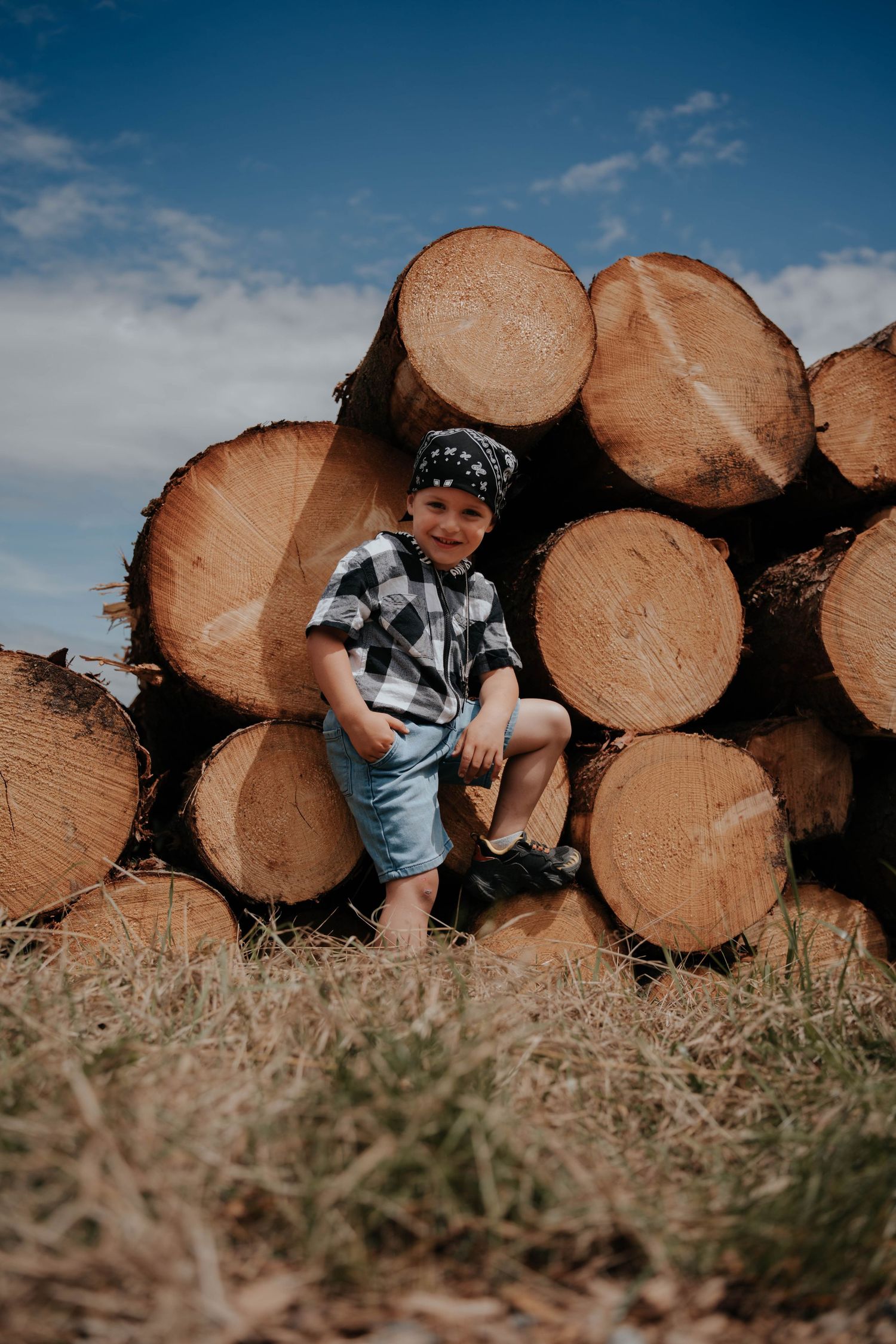 A smiling boy in a plaid shirt and bandana sits on a stack of cut logs under a blue sky.