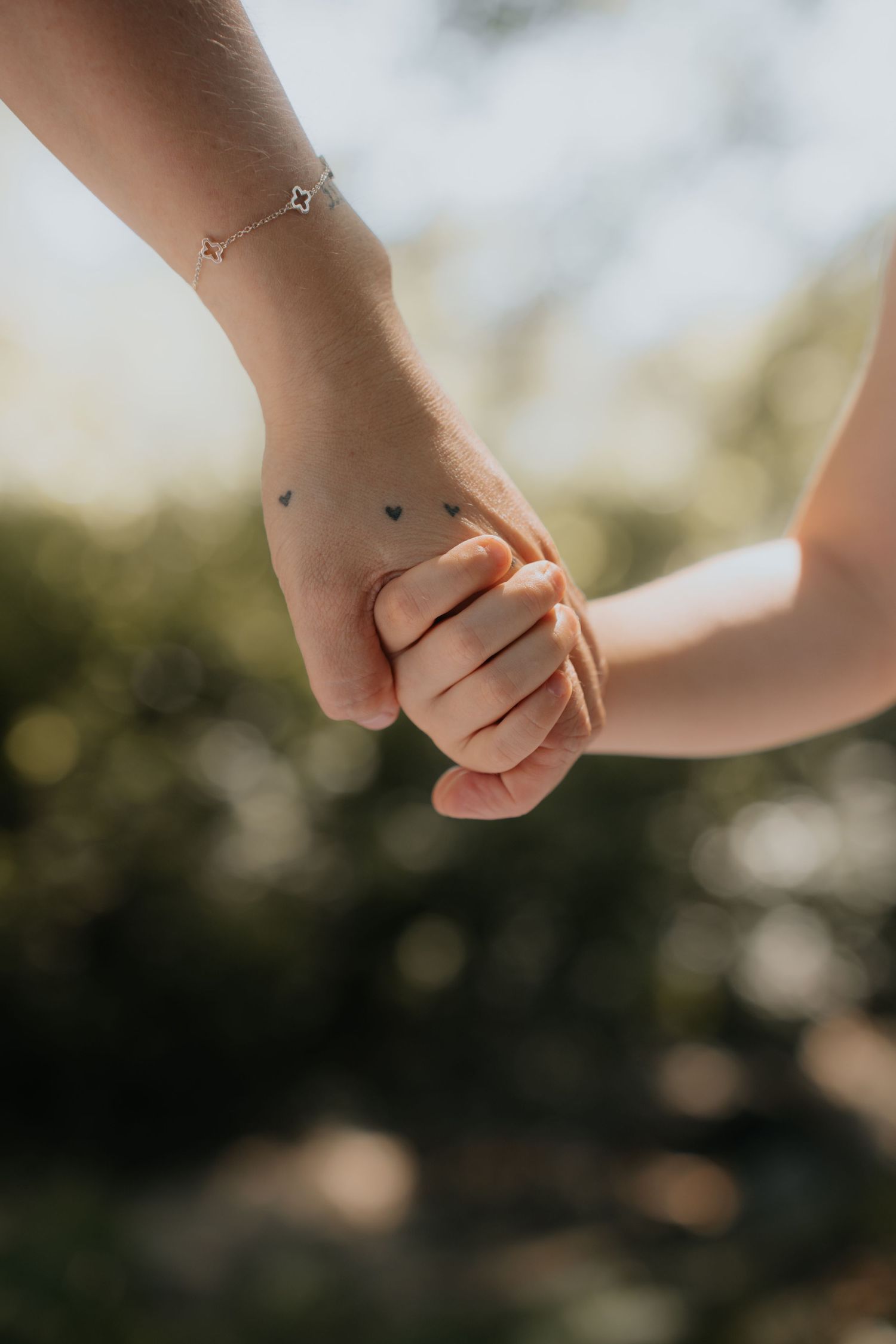 Adult hand with heart tattoos and star bracelet gently held by a small child's hand outdoors.