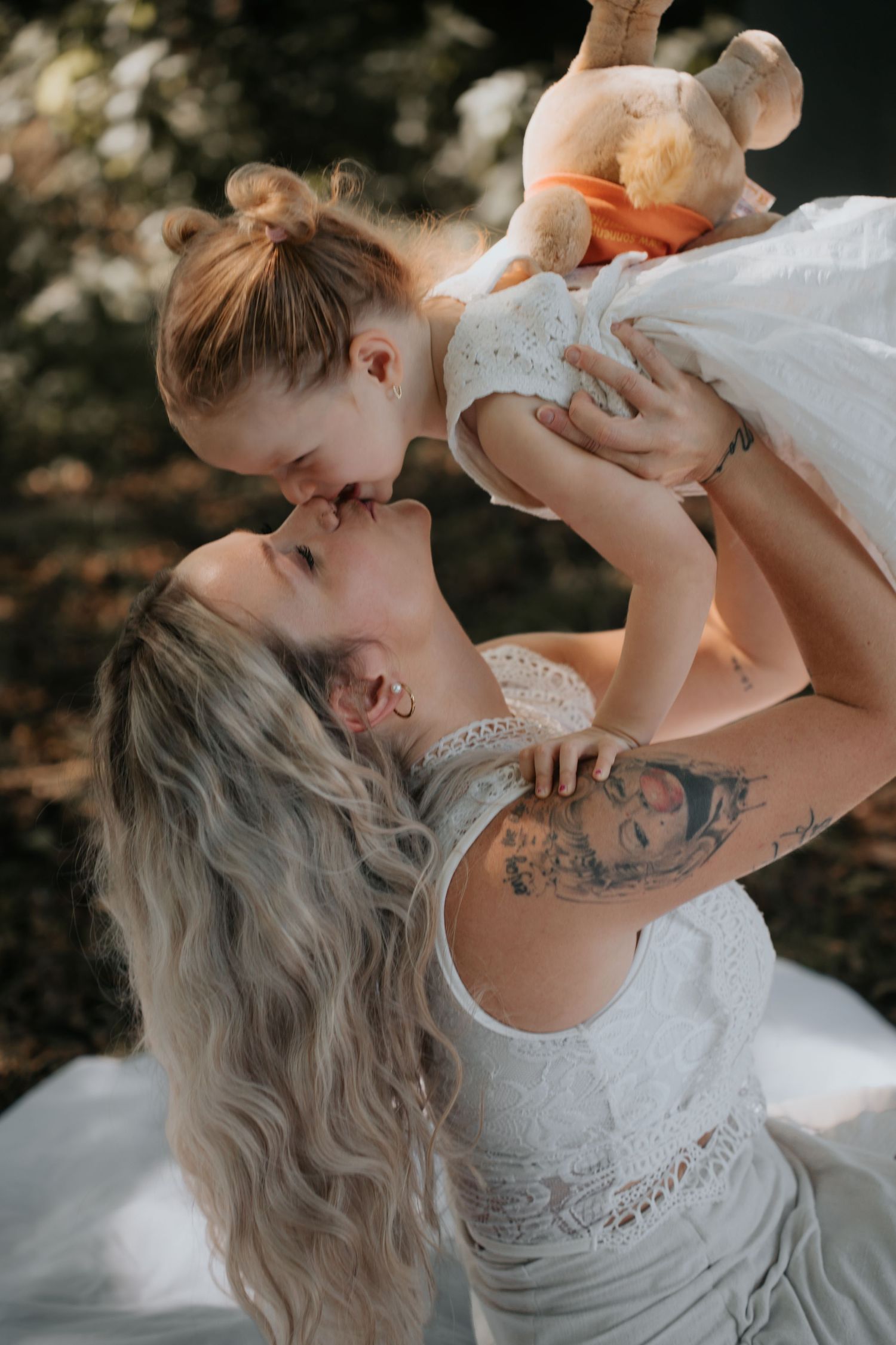 A tattooed mother lifts her toddler daughter for a kiss outdoors, both wearing white dresses.