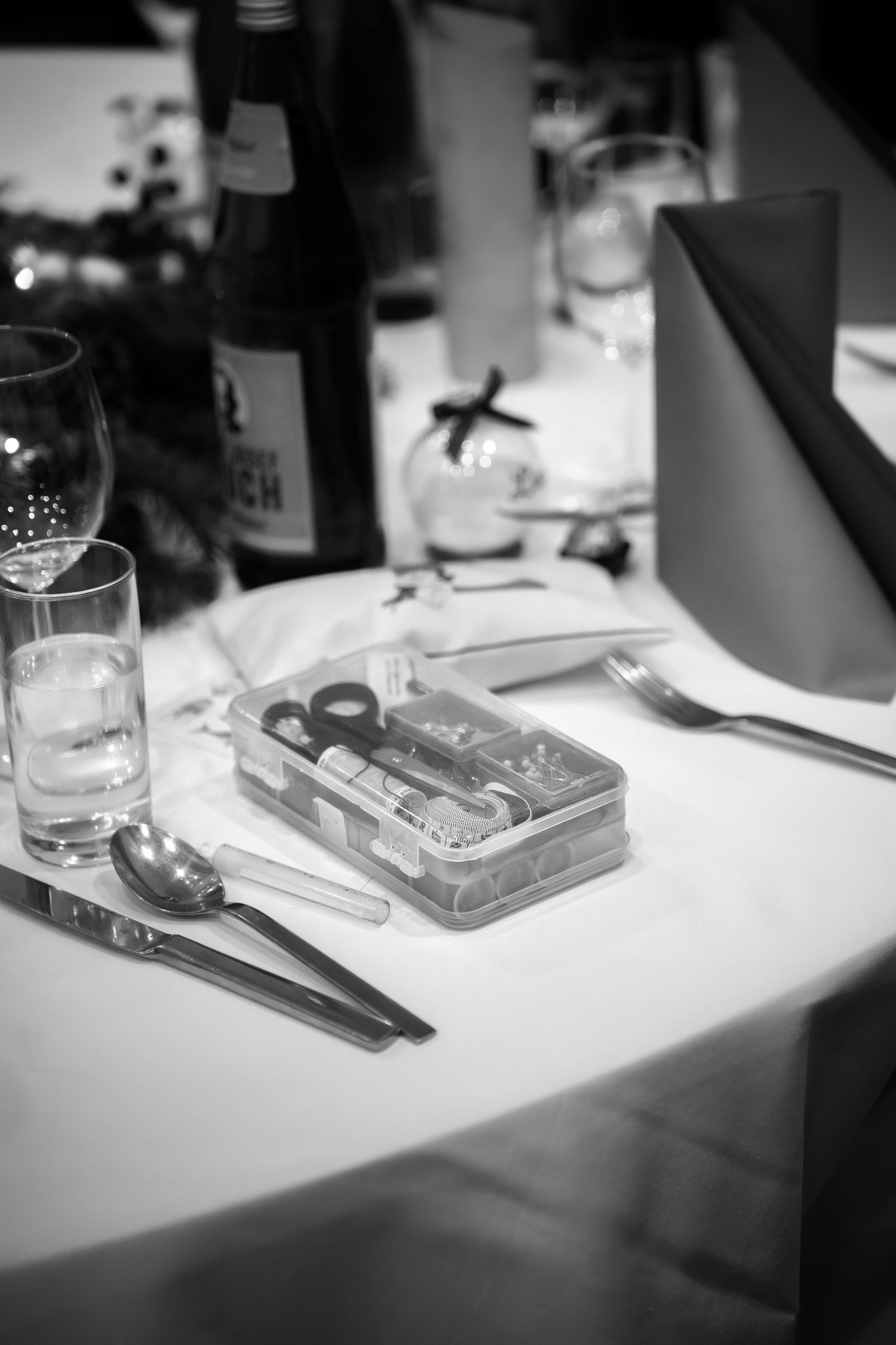 Black and white photo of an elegant table setting with cutlery, glassware, and a small kit box on white linen.