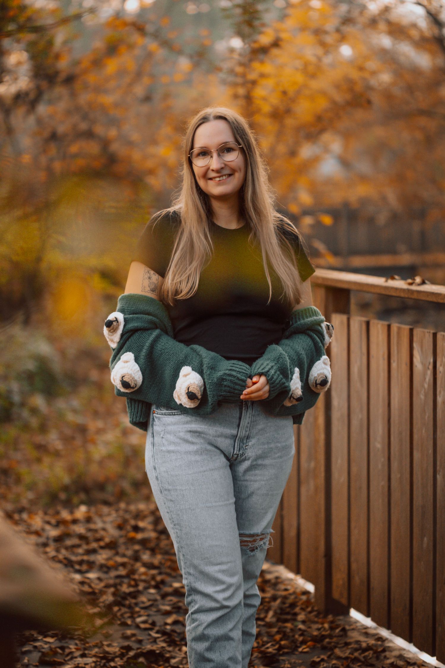 Smiling woman in green sweater and jeans holding a pug-themed cardigan outdoors in autumn foliage.