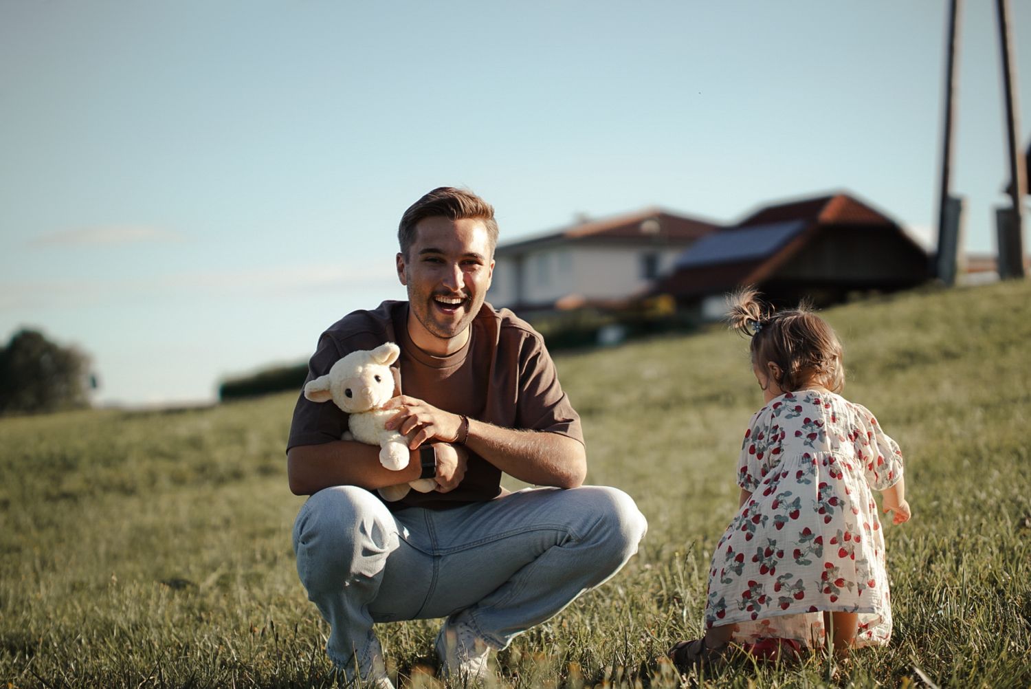 A smiling man crouches on green grass holding a teddy bear while a little girl in a floral dress plays nearby outdoors.