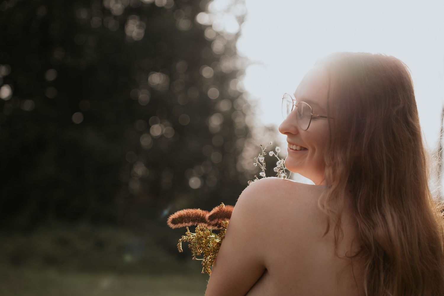 Smiling woman holding wildflowers in golden backlit sunlight outdoors.