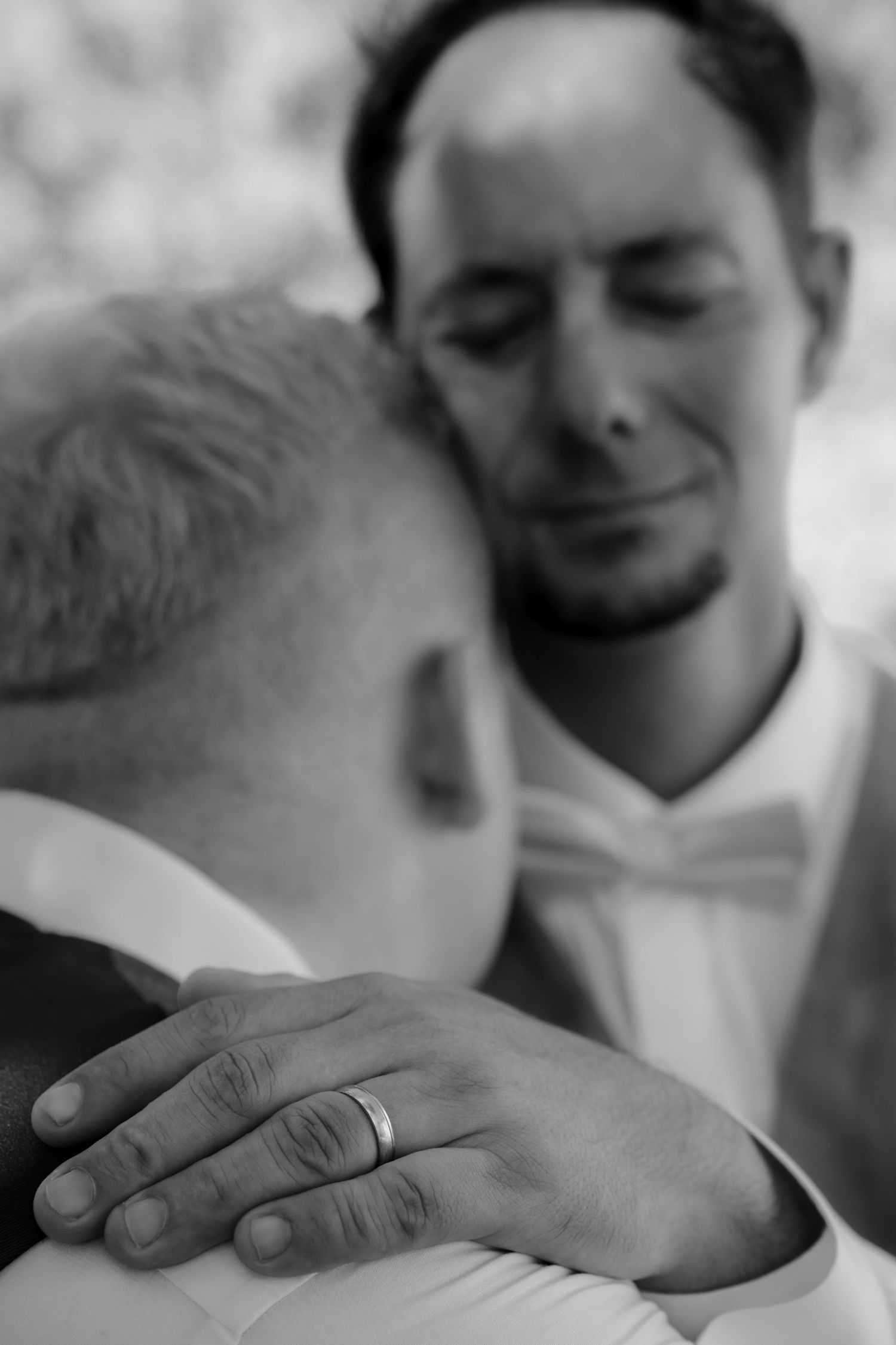 Black and white photo of two grooms embracing, one wearing a wedding ring on his hand resting on the other's shoulder.
