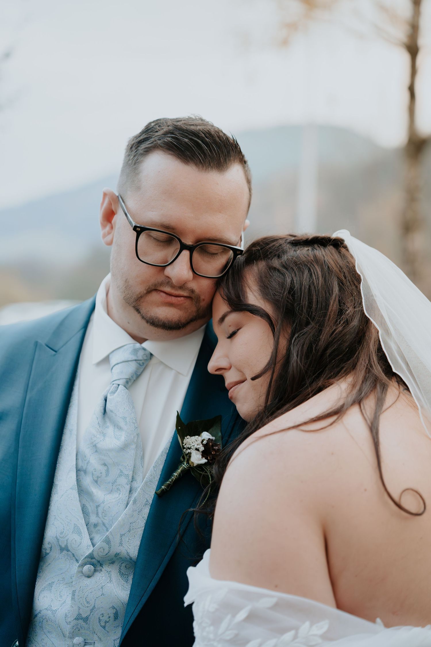 Bride and groom share tender moment, groom in blue suit with floral tie, bride in white veil, eyes closed outdoors.