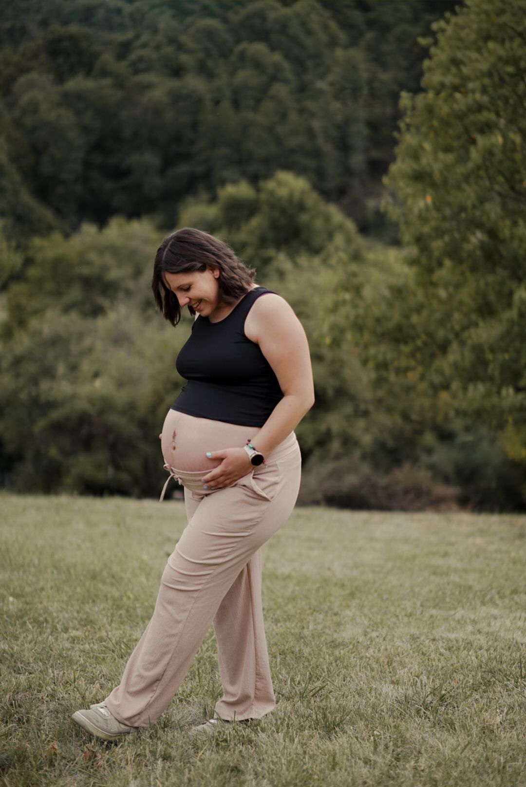 Pregnant woman in black crop top and beige pants cradling baby bump outdoors in green meadow.