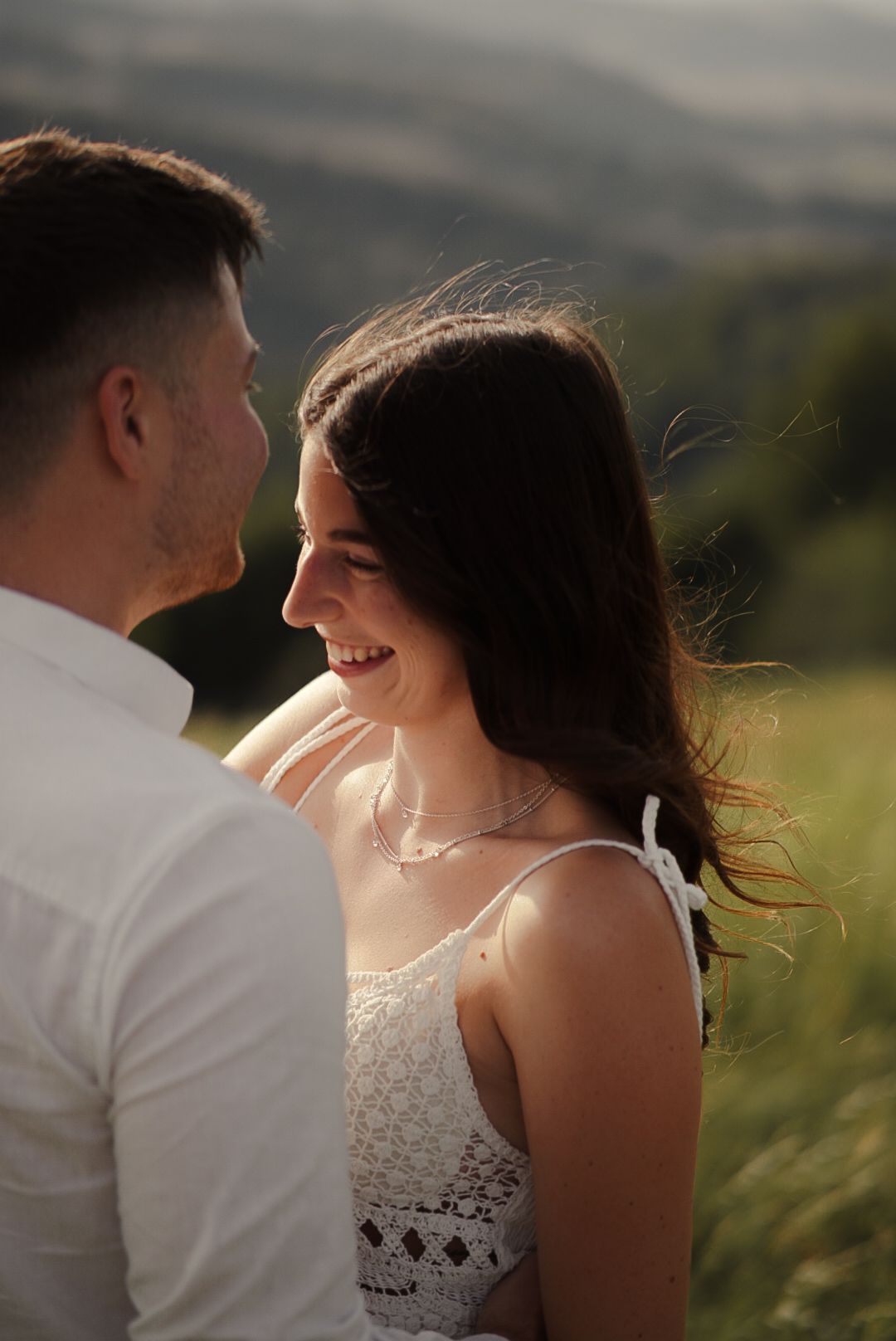 A smiling brunette woman in a white lace dress gazes at a man outdoors in golden sunlight.