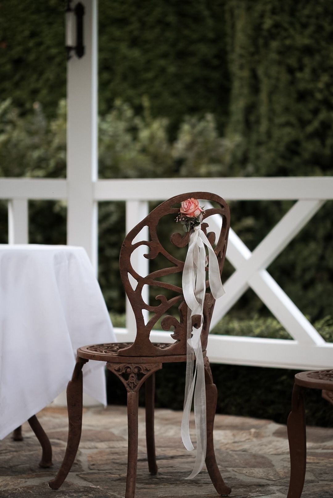 Ornate iron garden chair with pink rose and ribbon decoration on a stone patio near white fence.