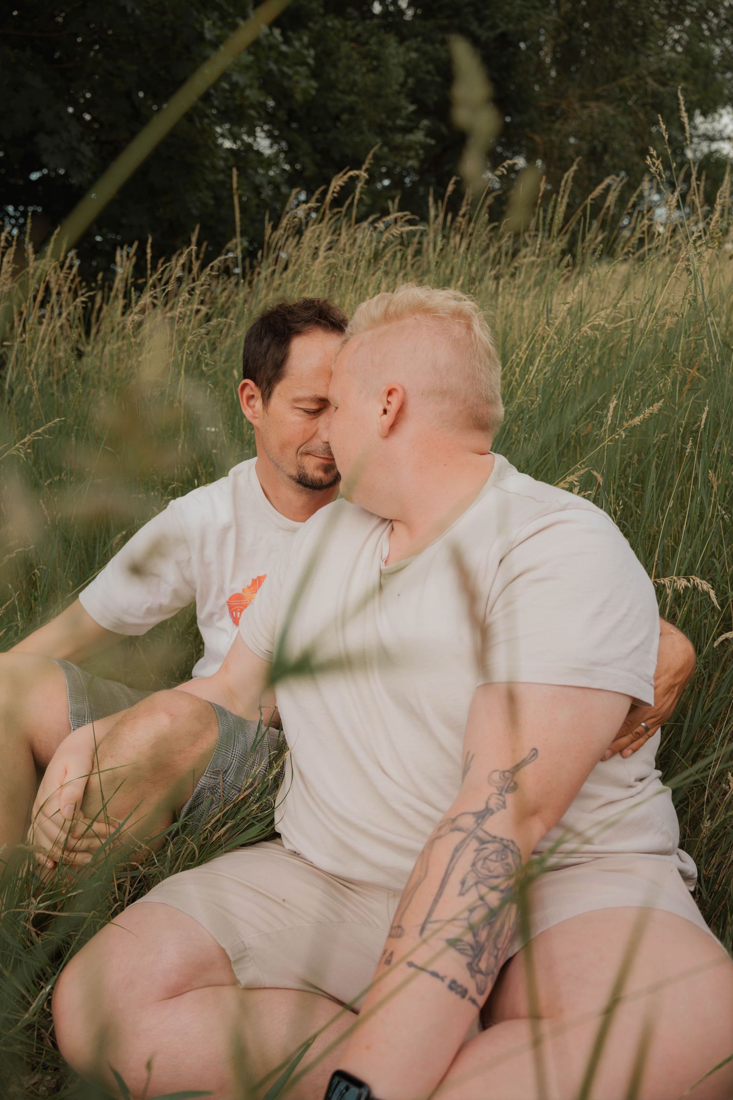 Two men sharing a romantic moment sitting in tall grass field, one with tattoos, both wearing white t-shirts outdoors.