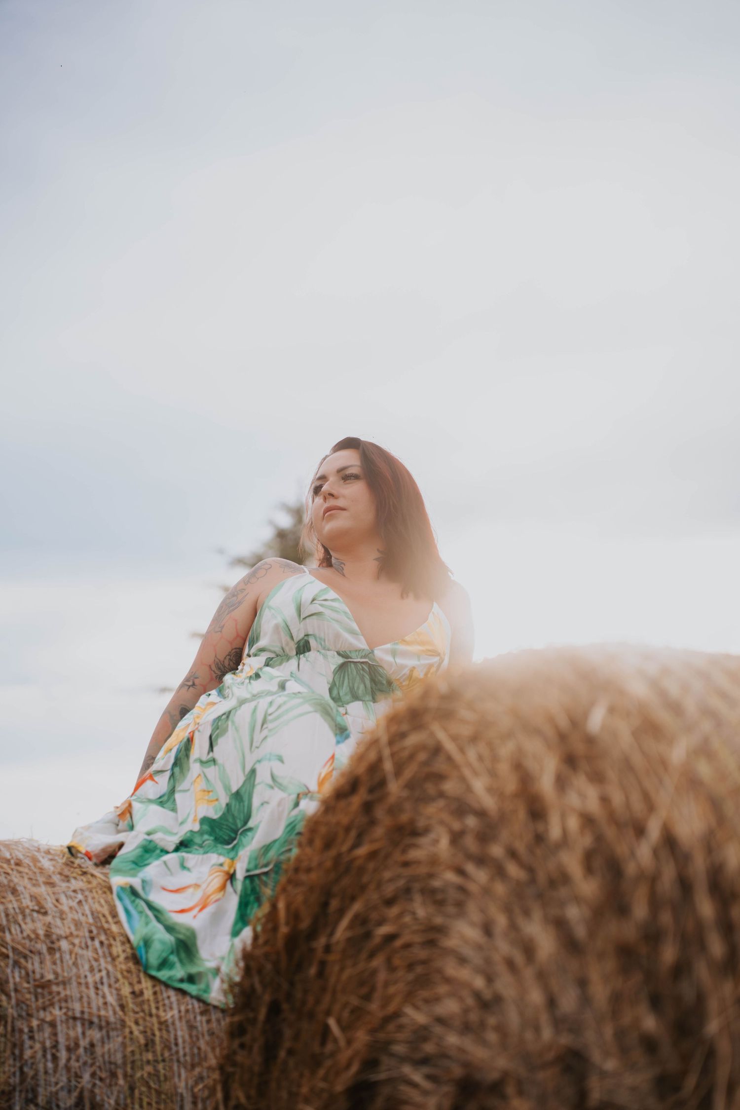 Woman in floral dress sitting atop a hay bale in a field, gazing into the distance under a cloudy sky.