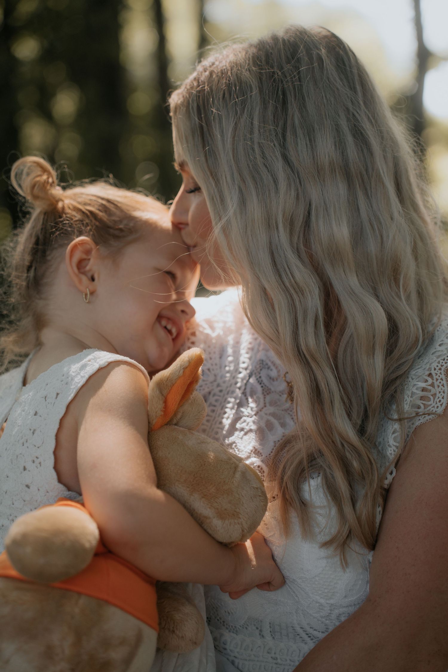 A mother with blonde hair kisses her laughing toddler daughter holding a stuffed animal outdoors in warm sunlight.