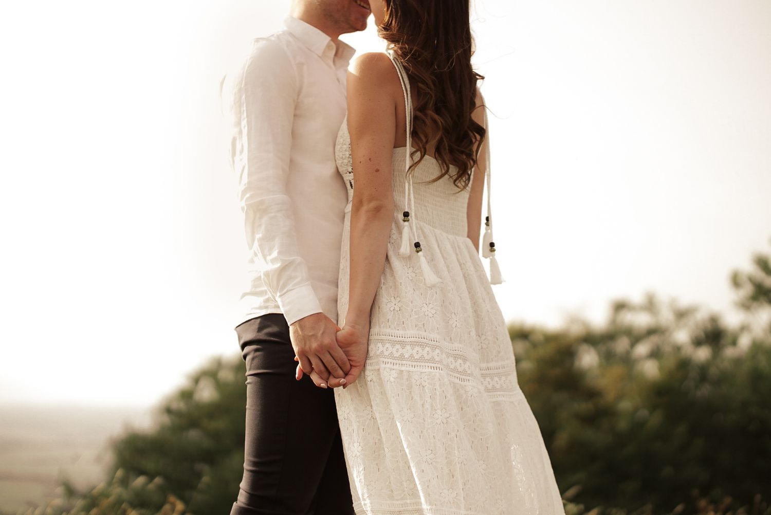 Romantic couple holding hands outdoors, woman in white lace dress and man in white shirt, soft natural background.