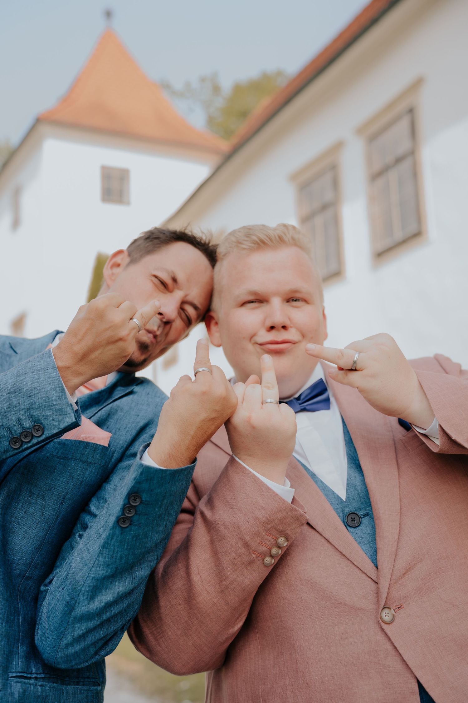 Two grooms in suits showing wedding rings, posing playfully outside a white building on their wedding day.
