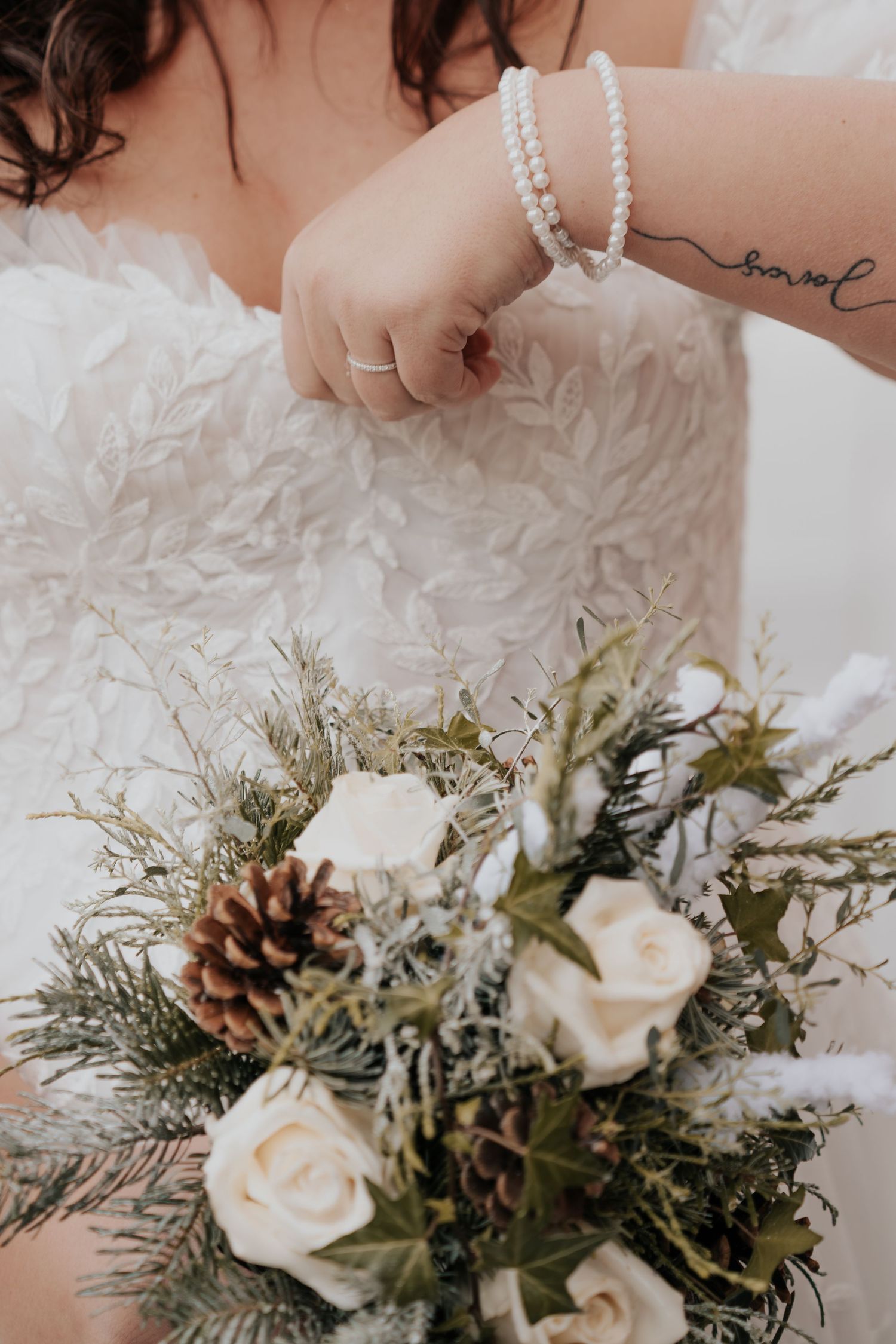Bride in white lace gown holding winter bouquet with roses, pine cones, and greenery, wearing pearl bracelet.