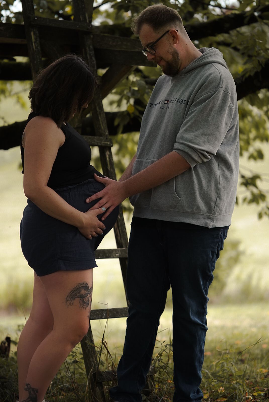 A couple shares a tender maternity moment outdoors, with the man gently touching the pregnant woman's baby bump near a rustic wooden fence.