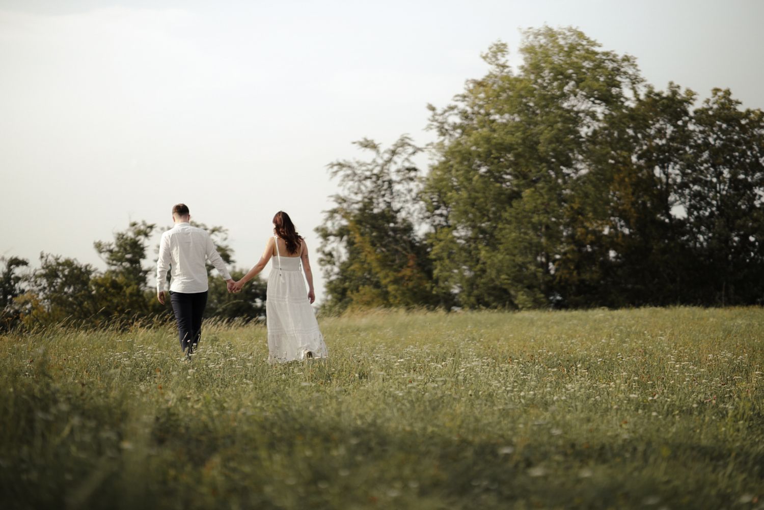 A couple walks hand-in-hand through a lush green meadow, the bride in a white dress, surrounded by trees.