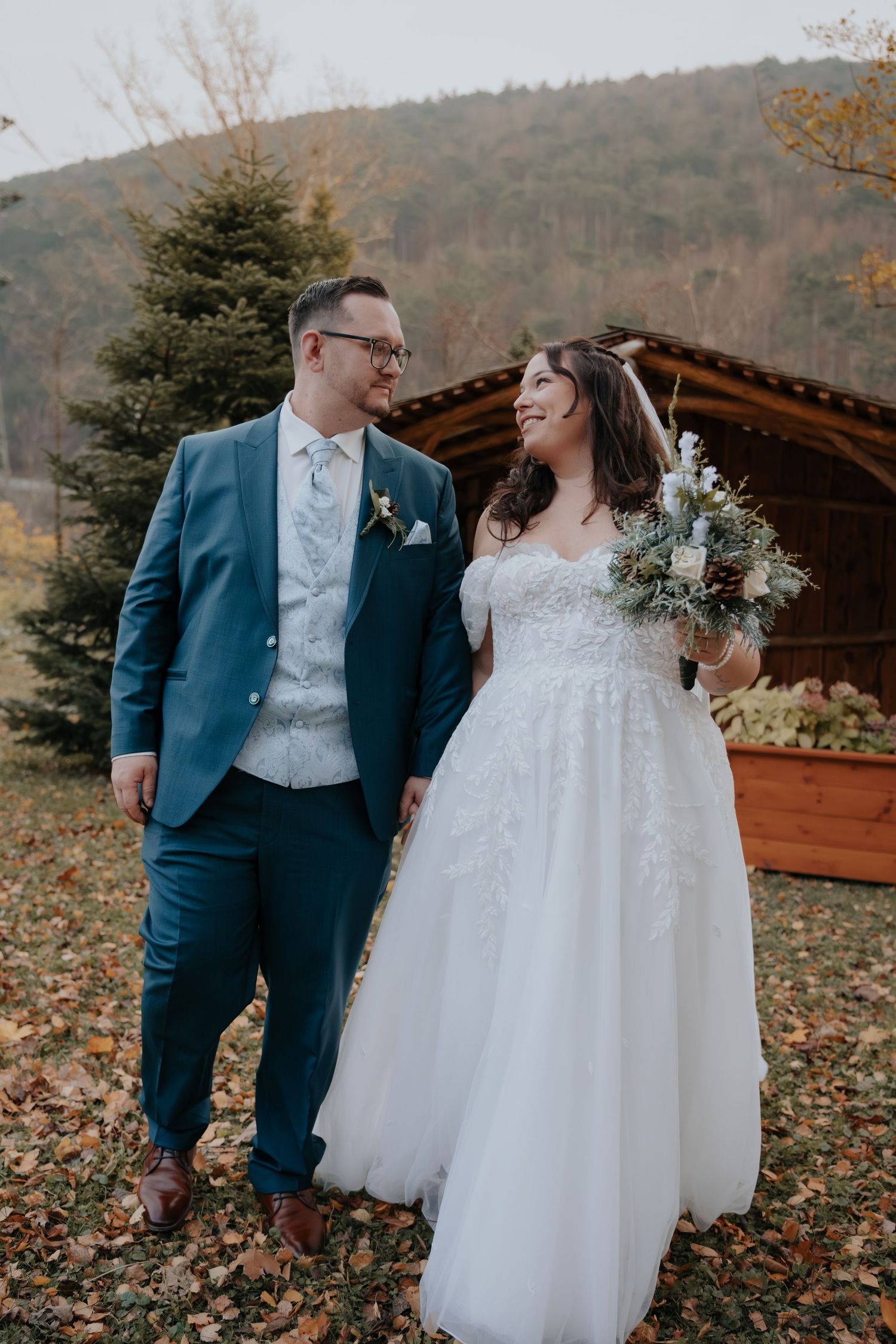 Bride and groom pose outdoors in autumn, she holds a bouquet in a white gown, he wears a blue suit.