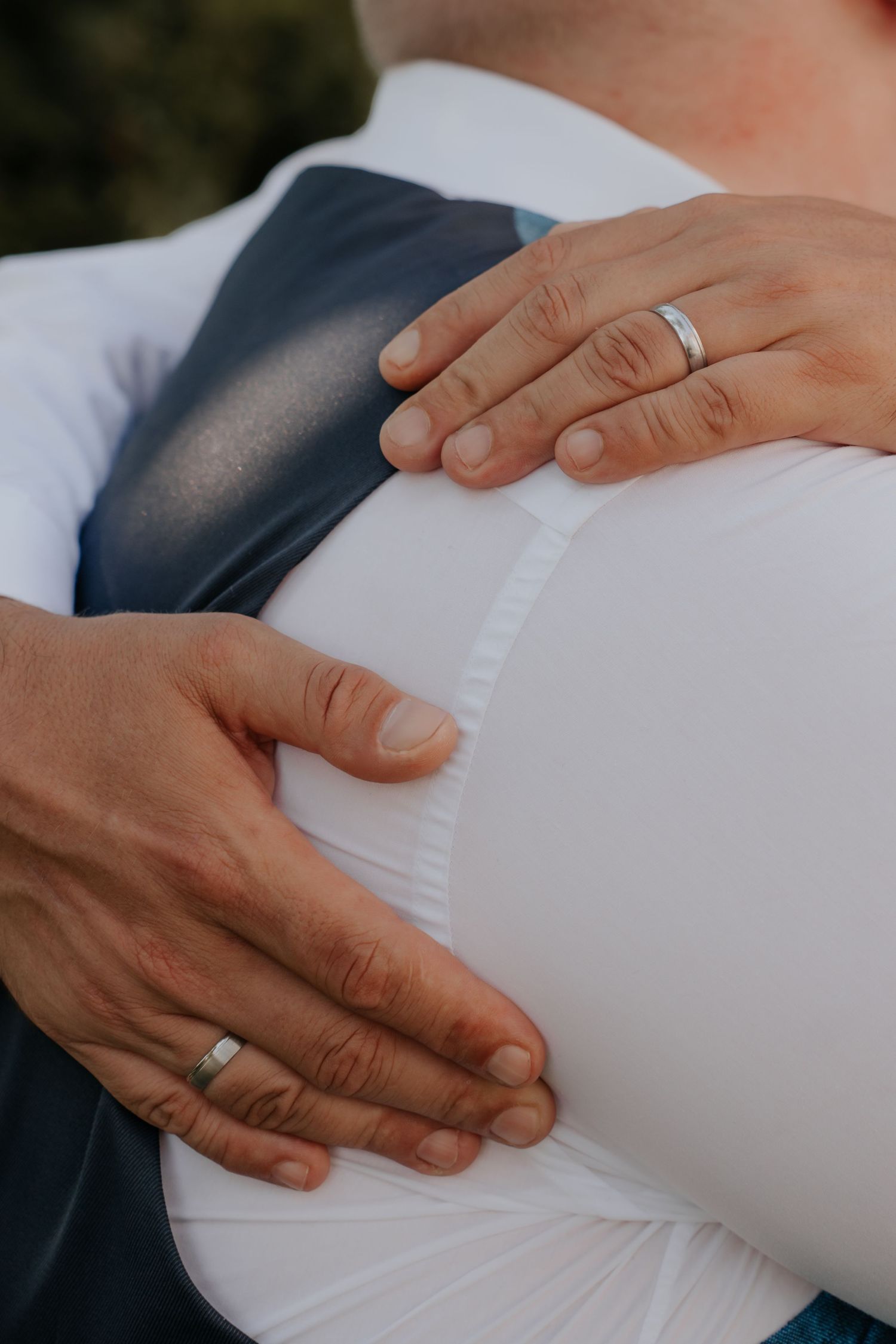 Close-up of two hands with wedding rings tenderly cradling a pregnant belly in a white shirt.