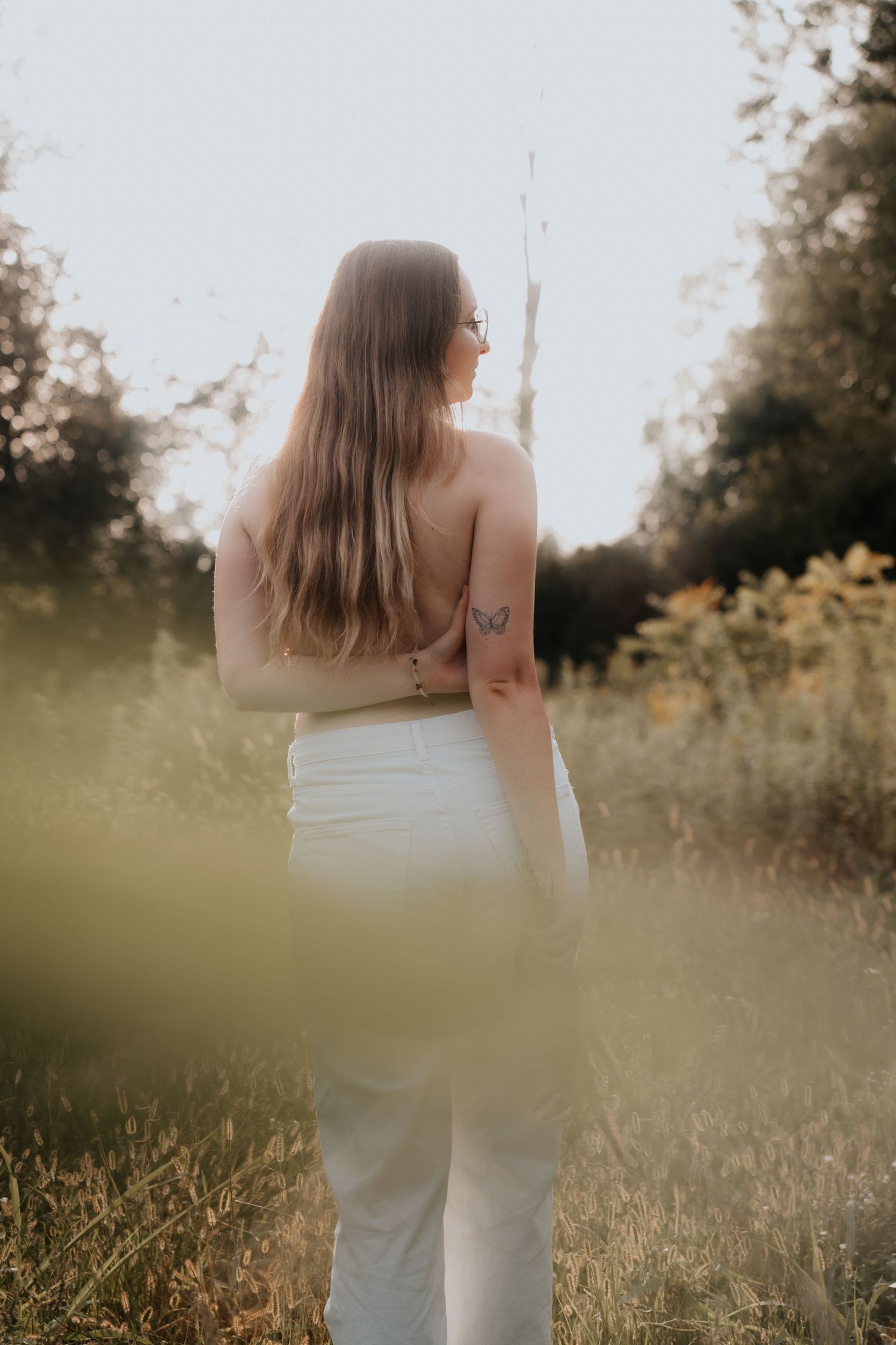 Woman with long hair and butterfly tattoo stands in golden field wearing white pants, viewed from behind at sunset.