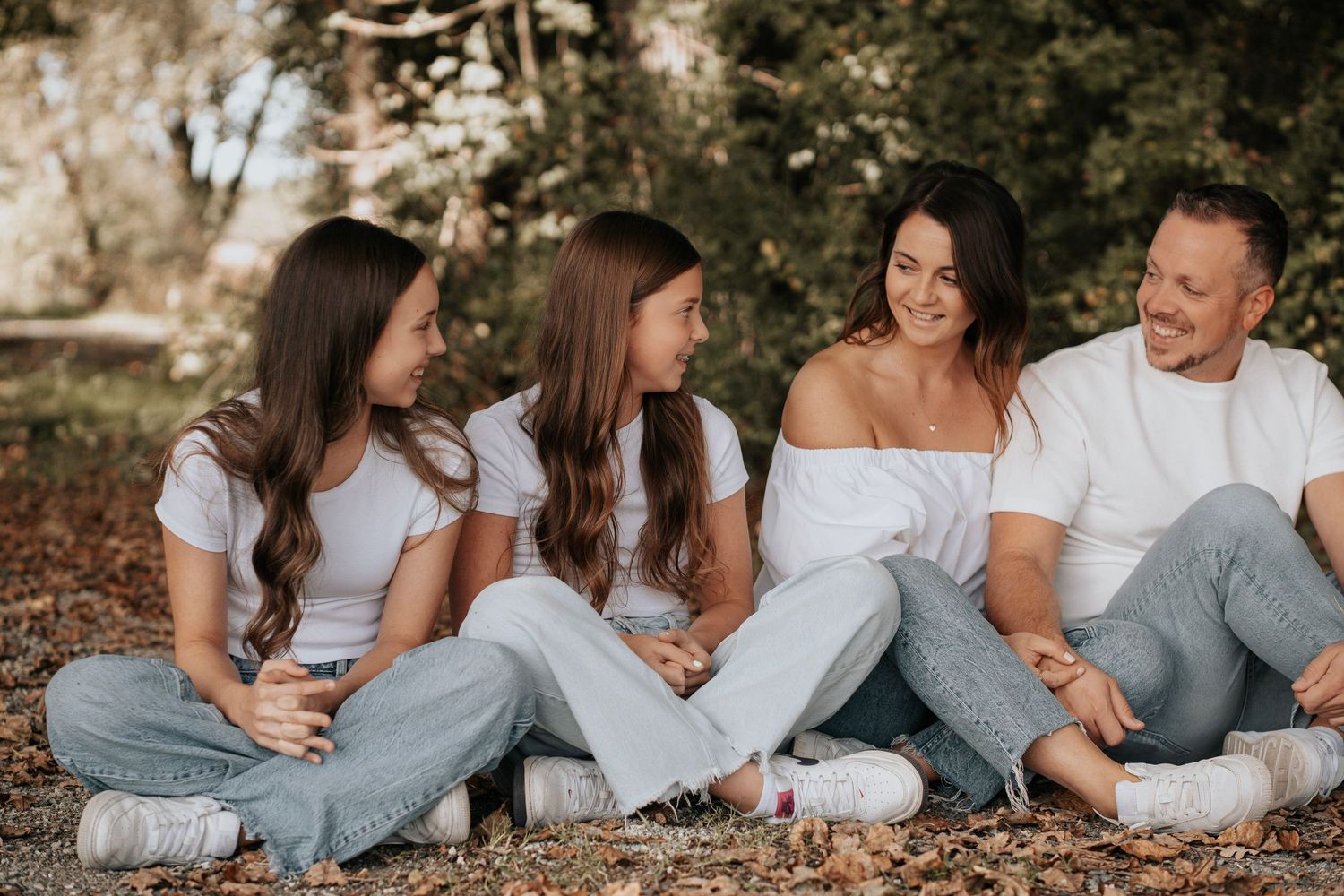 Family of four in white tops and jeans sitting outdoors on ground, smiling and laughing together in a sunny park.