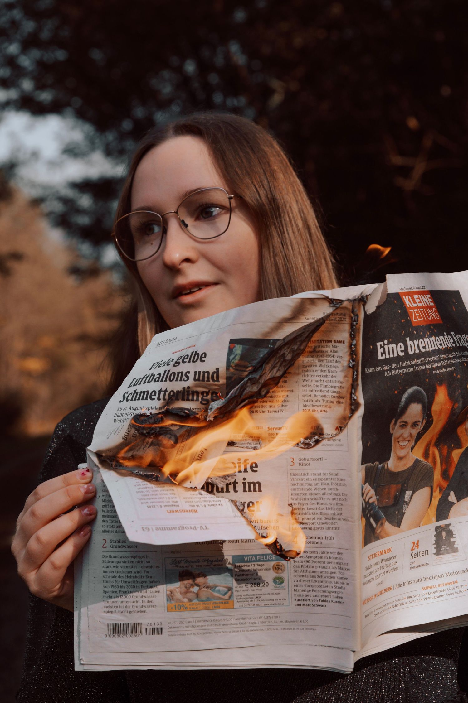 Woman with glasses holding a burning newspaper outdoors, flames consuming the pages of a German publication.
