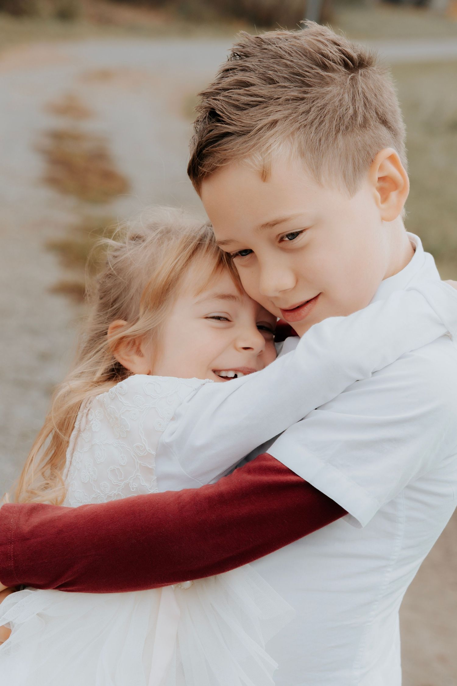Two young siblings share a sweet embrace outdoors, with a smiling girl in white hugging her older brother in a white shirt.