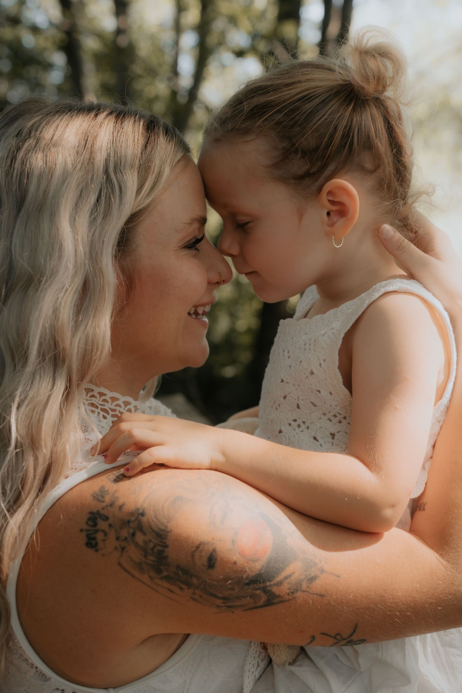 A tattooed mother tenderly holds her young daughter, touching foreheads in a loving outdoor moment.