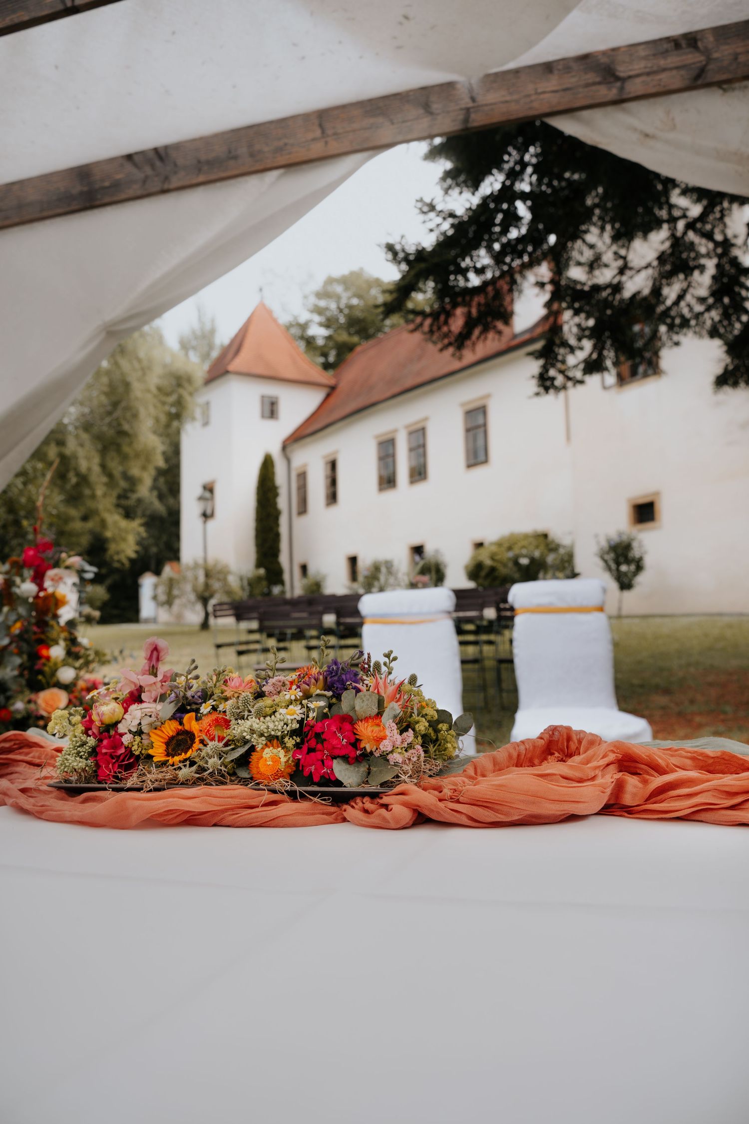 Colorful floral centerpiece on orange draped table at outdoor wedding venue with white manor house in background.