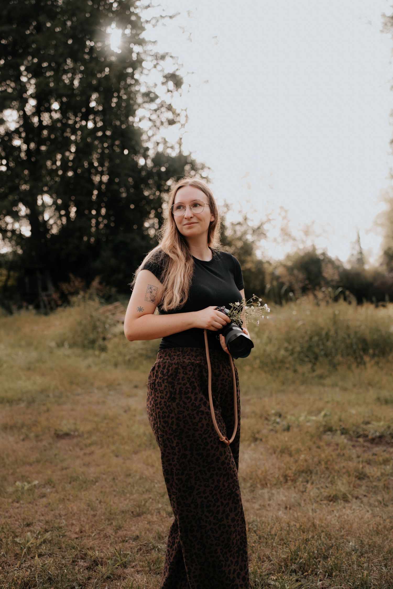Woman holding camera and wildflowers in a sunlit field, wearing black top and floral skirt, golden hour photography.