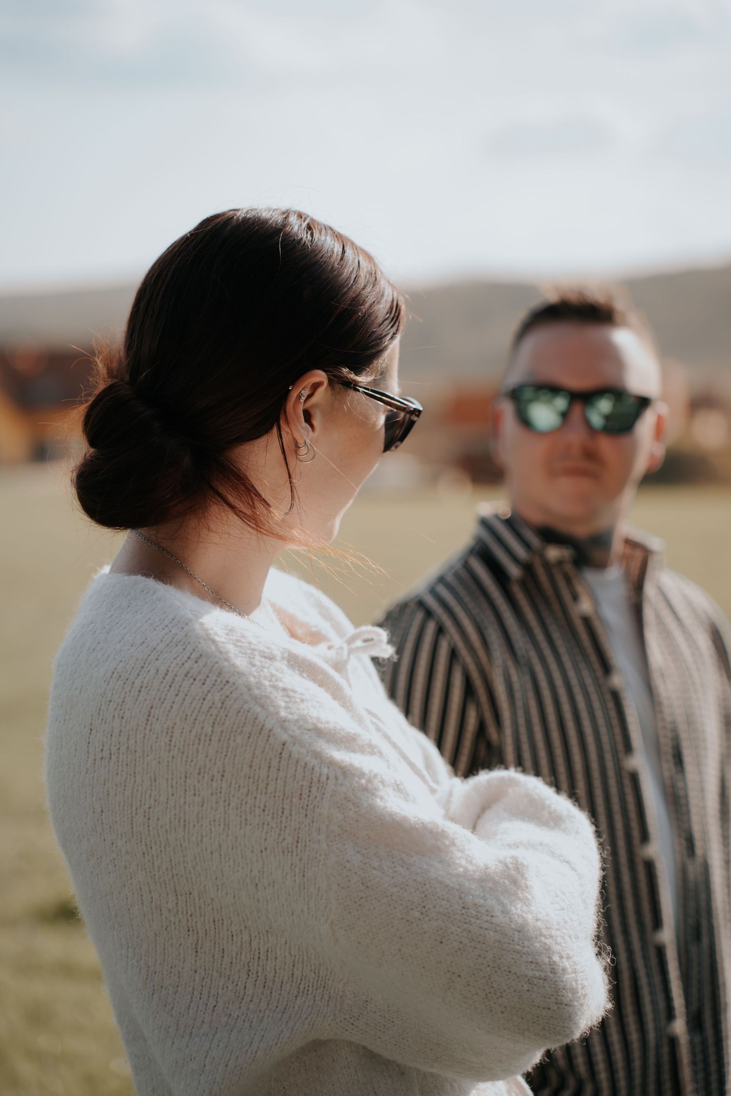 Young couple outdoors, woman in white sweater and sunglasses with arms crossed, man in striped shirt behind her.