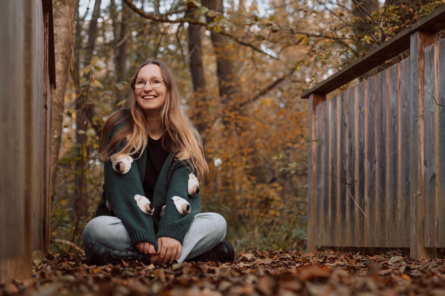 Smiling woman sitting cross-legged on autumn leaves outdoors, wearing a green sweater and jeans near wooden structures.