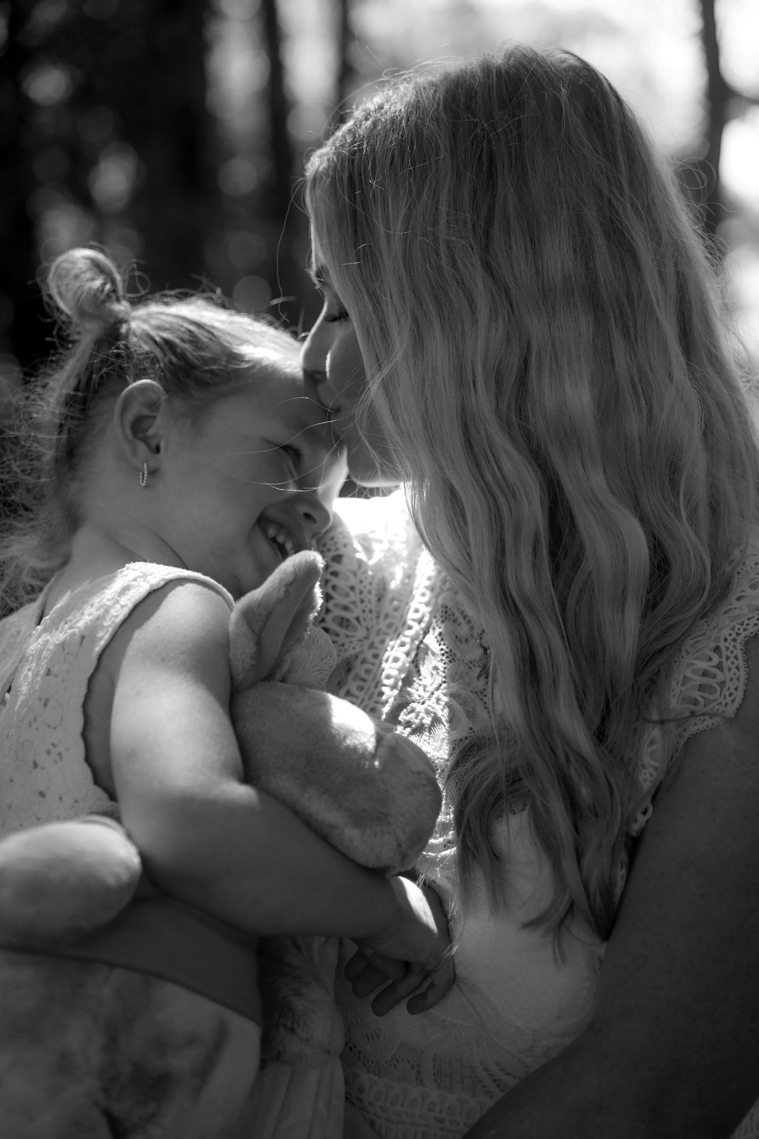 Black and white photo of a mother and young daughter smiling at each other, girl holding a stuffed animal.