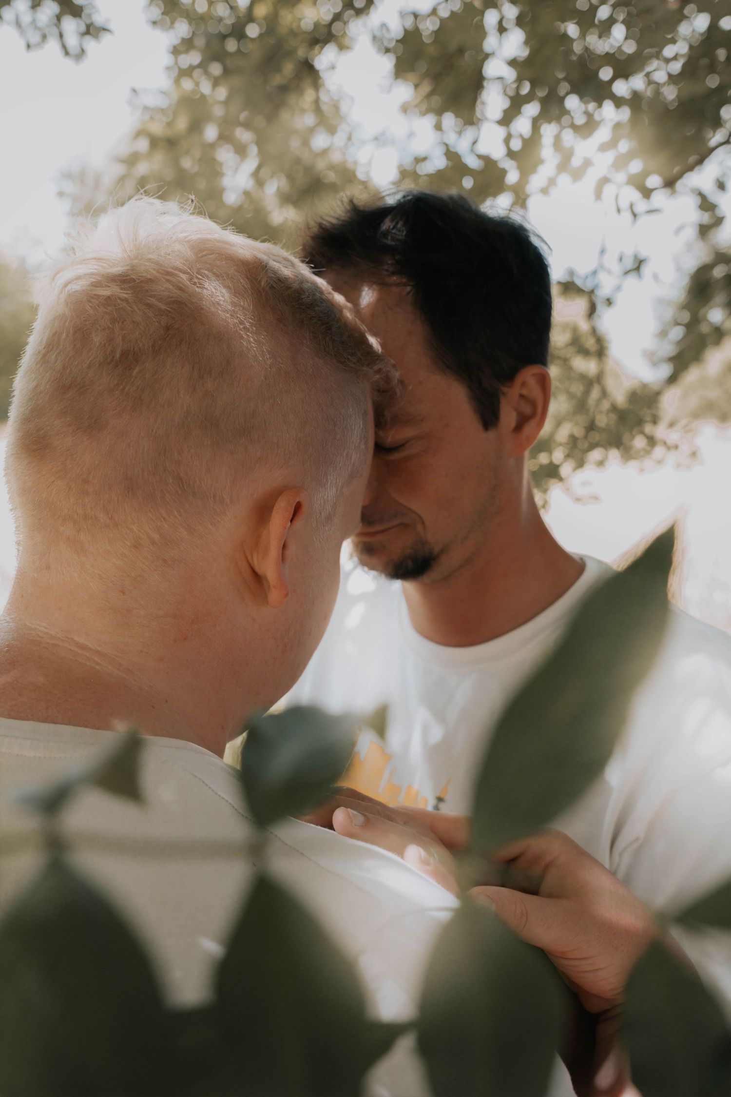 Two men share an intimate moment outdoors, surrounded by soft bokeh greenery in warm natural light.